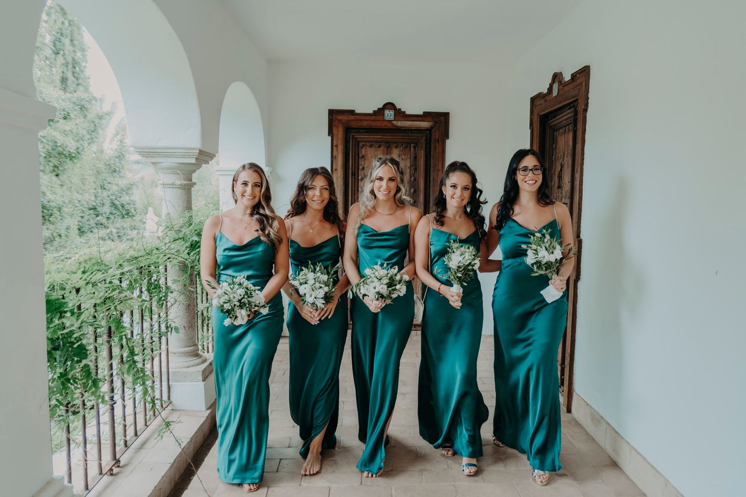 Bridesmaids in matching teal satin dresses holding white bouquets pose together on a covered balcony.