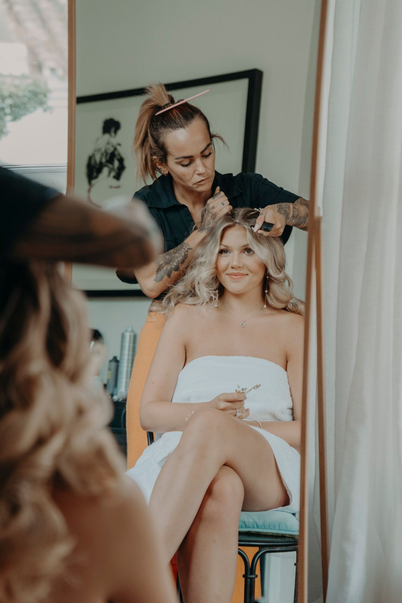 A hairstylist works on styling a bride's blonde hair as she sits in a white wrap during wedding preparations.