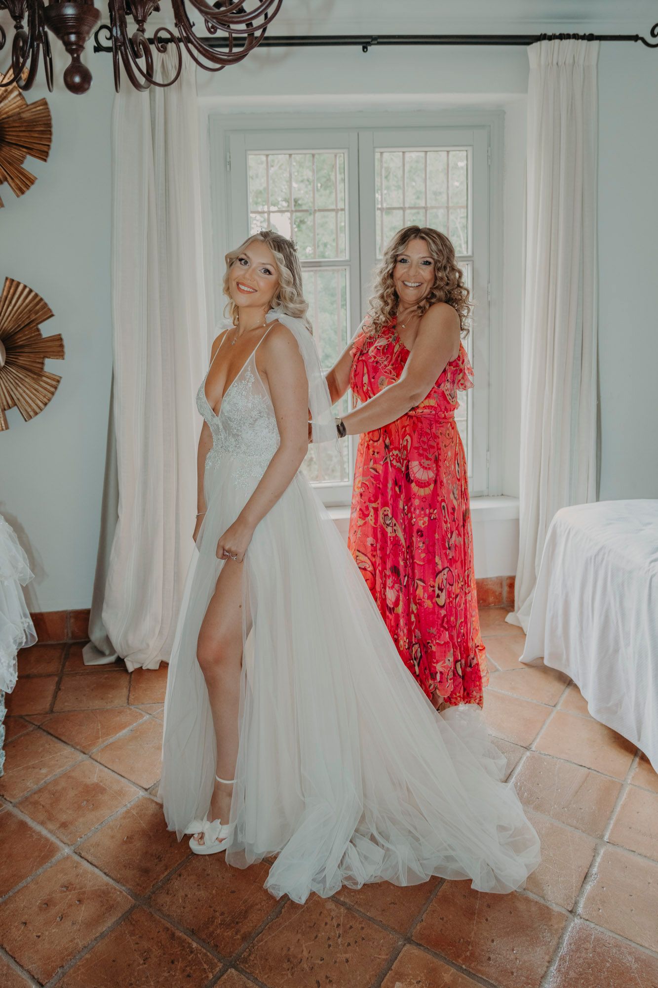 Two women posing in elegant dresses, one wearing a white wedding gown and one in a flowing red floral dress.