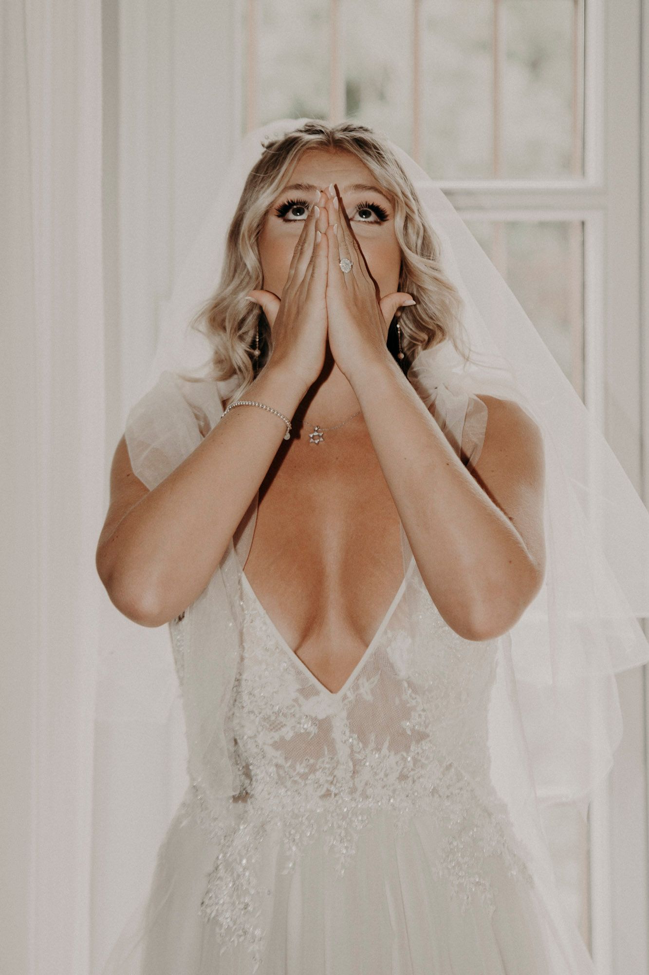 Woman in white wedding dress and veil covers her face with her hands in an emotional moment.