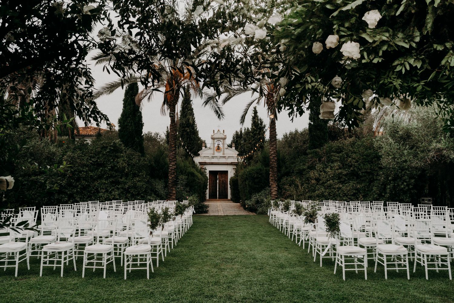 Outdoor wedding ceremony setup with rows of white chairs leading to a gazebo under cypress trees.