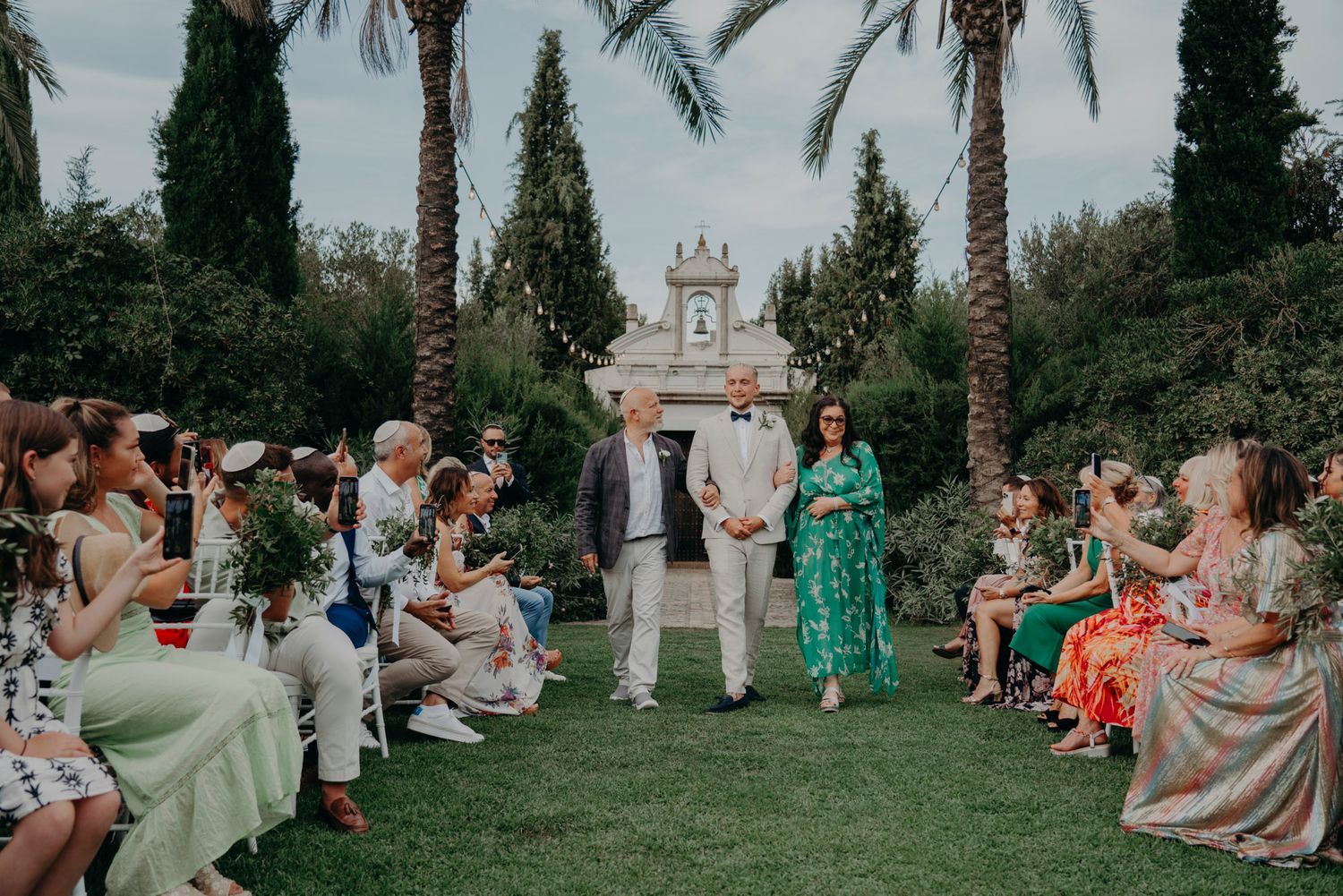 Outdoor garden wedding ceremony with guests seated on lawn chairs amid palm trees and cypress trees.