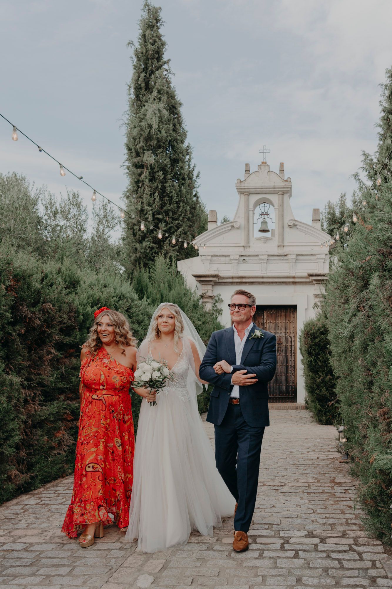Outside an elegant chapel with cypress trees, a bride in white walks with two guests down a stone pathway.