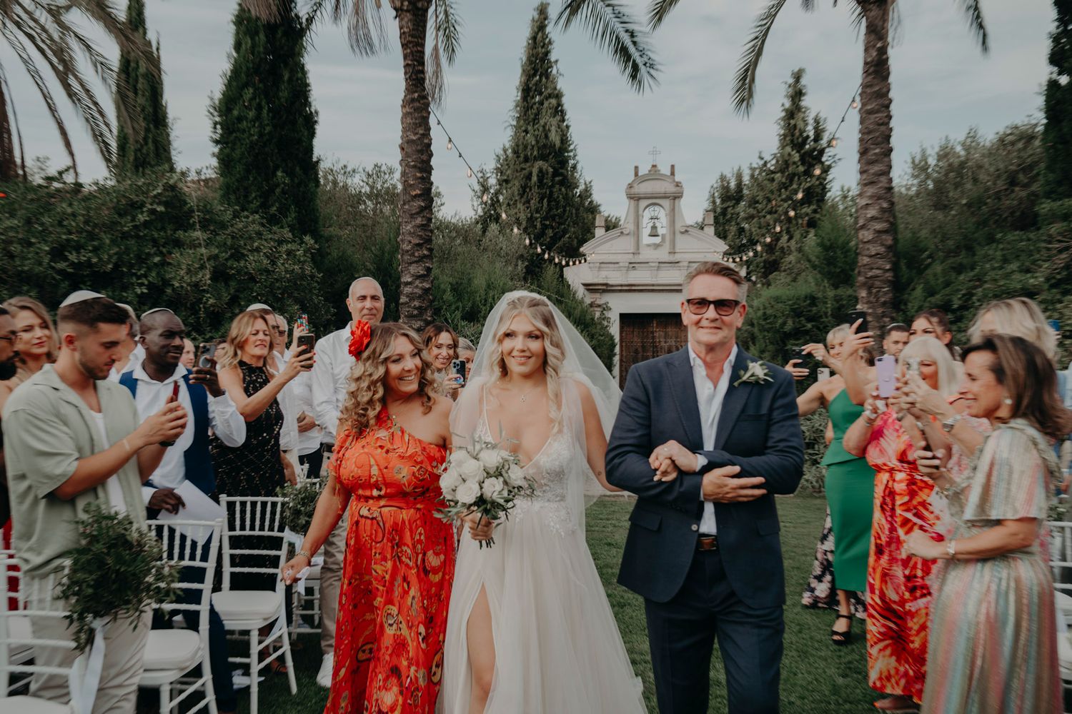 Wedding ceremony guests lined up outdoors with palm trees and a white architectural structure in the background.