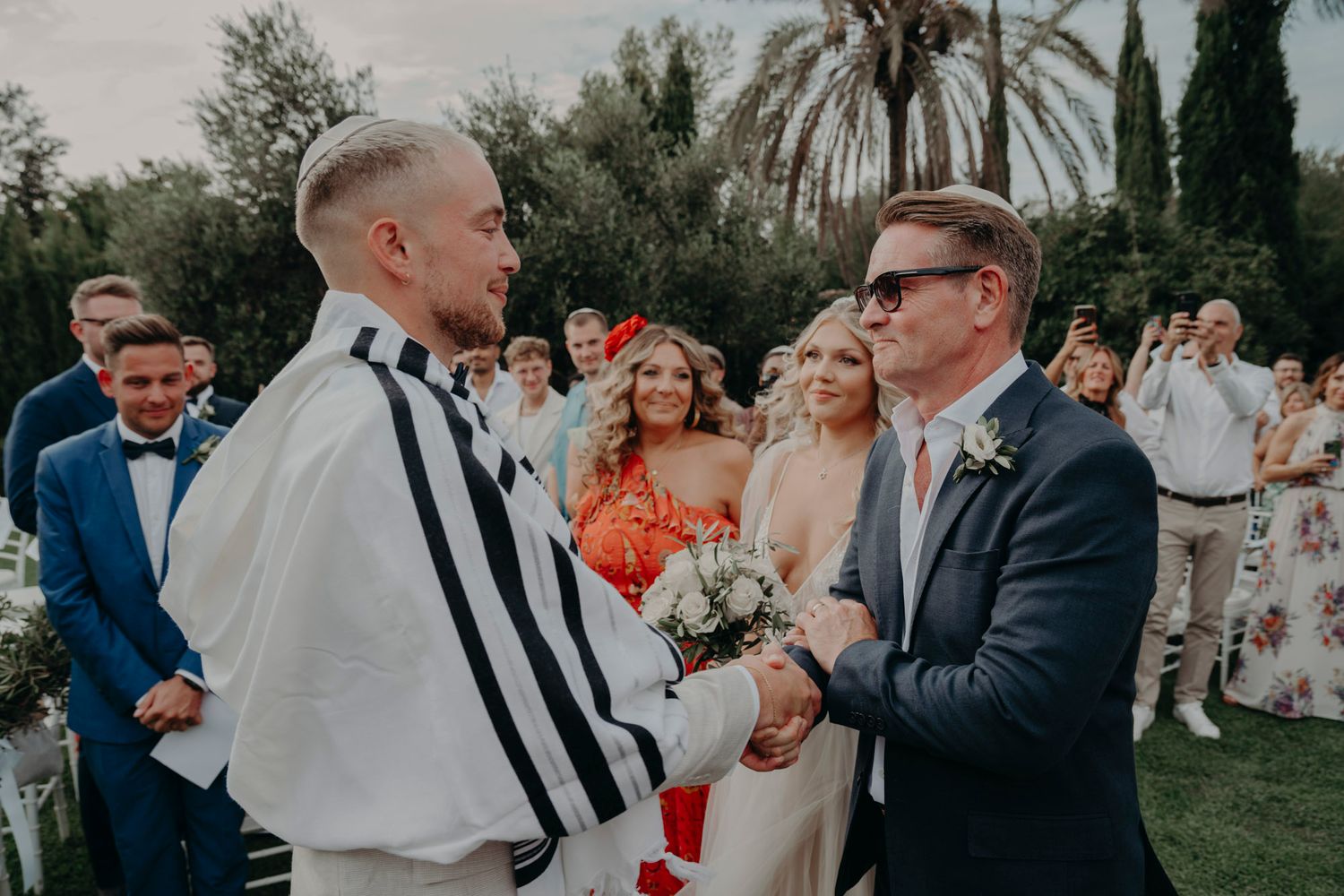 Wedding guests gather in a garden setting with palm trees and cypress trees in the background during an outdoor celebration.