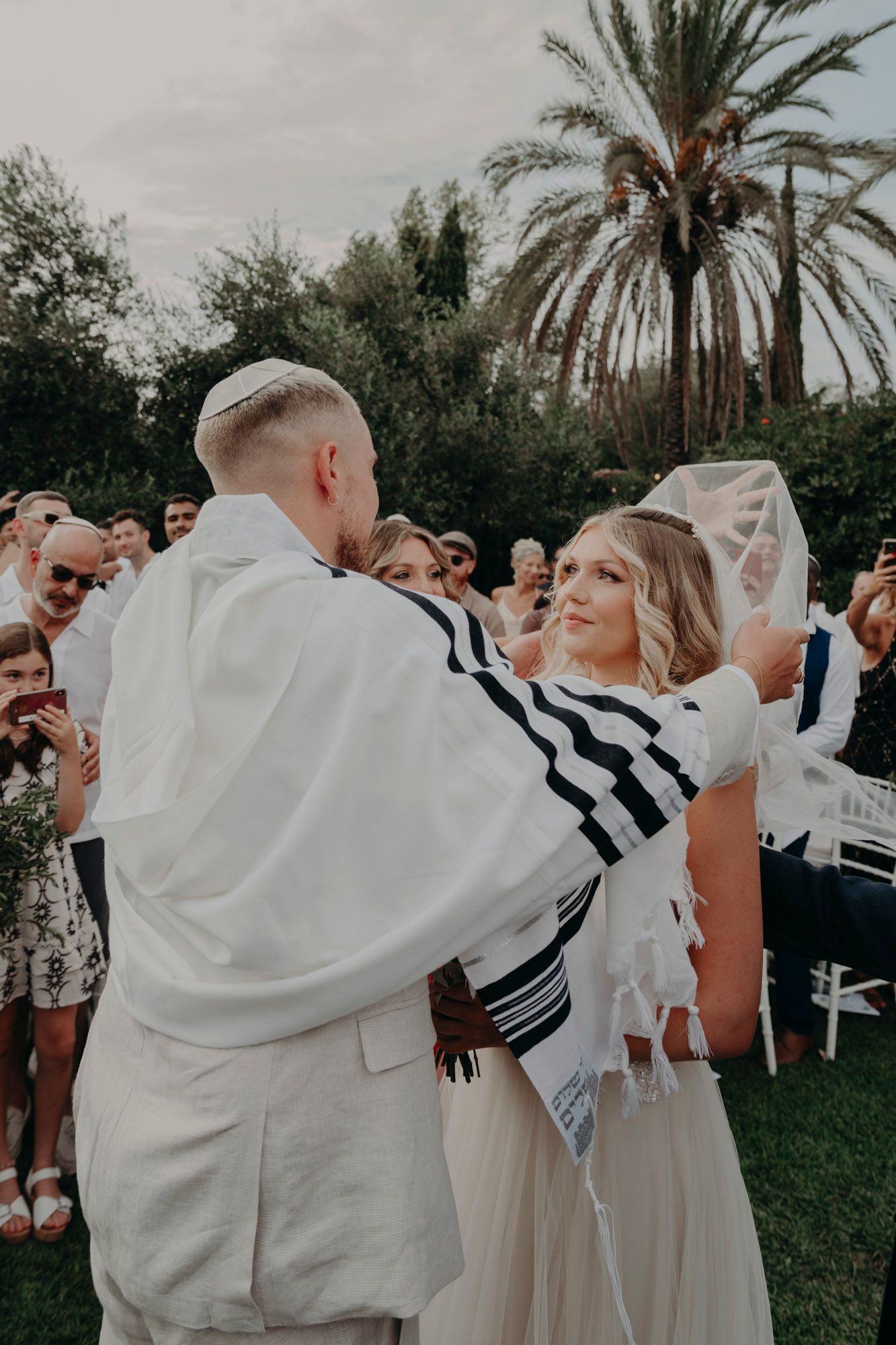 A wedding celebration takes place outdoors with palm trees in the background during a traditional Jewish ceremony.