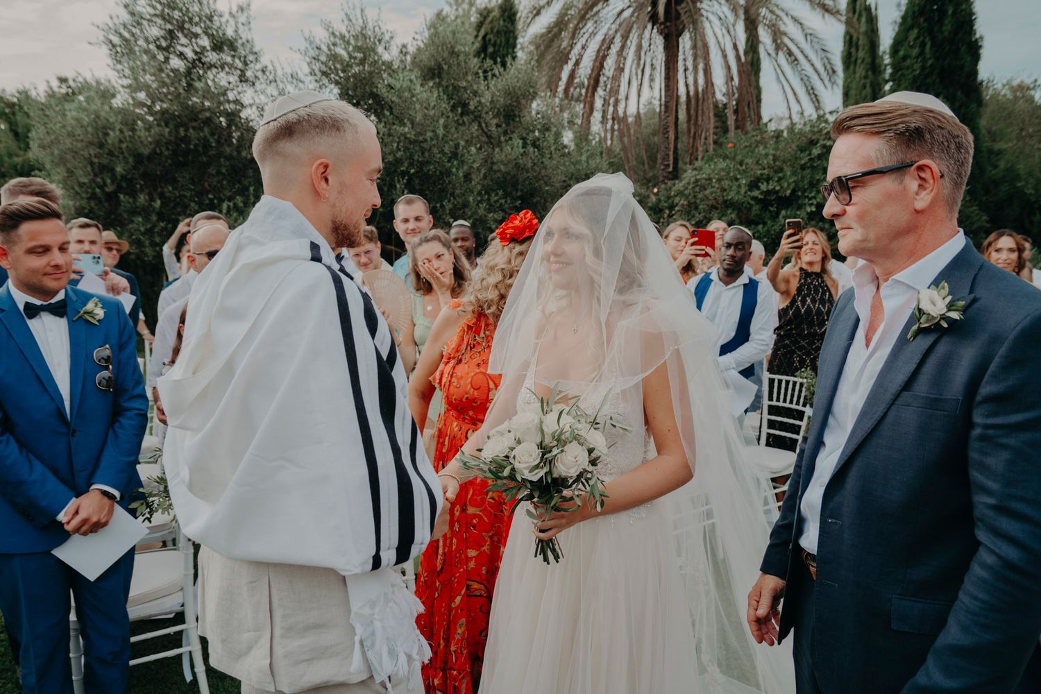 Outdoor wedding ceremony with guests gathered around in formal attire under palm trees.