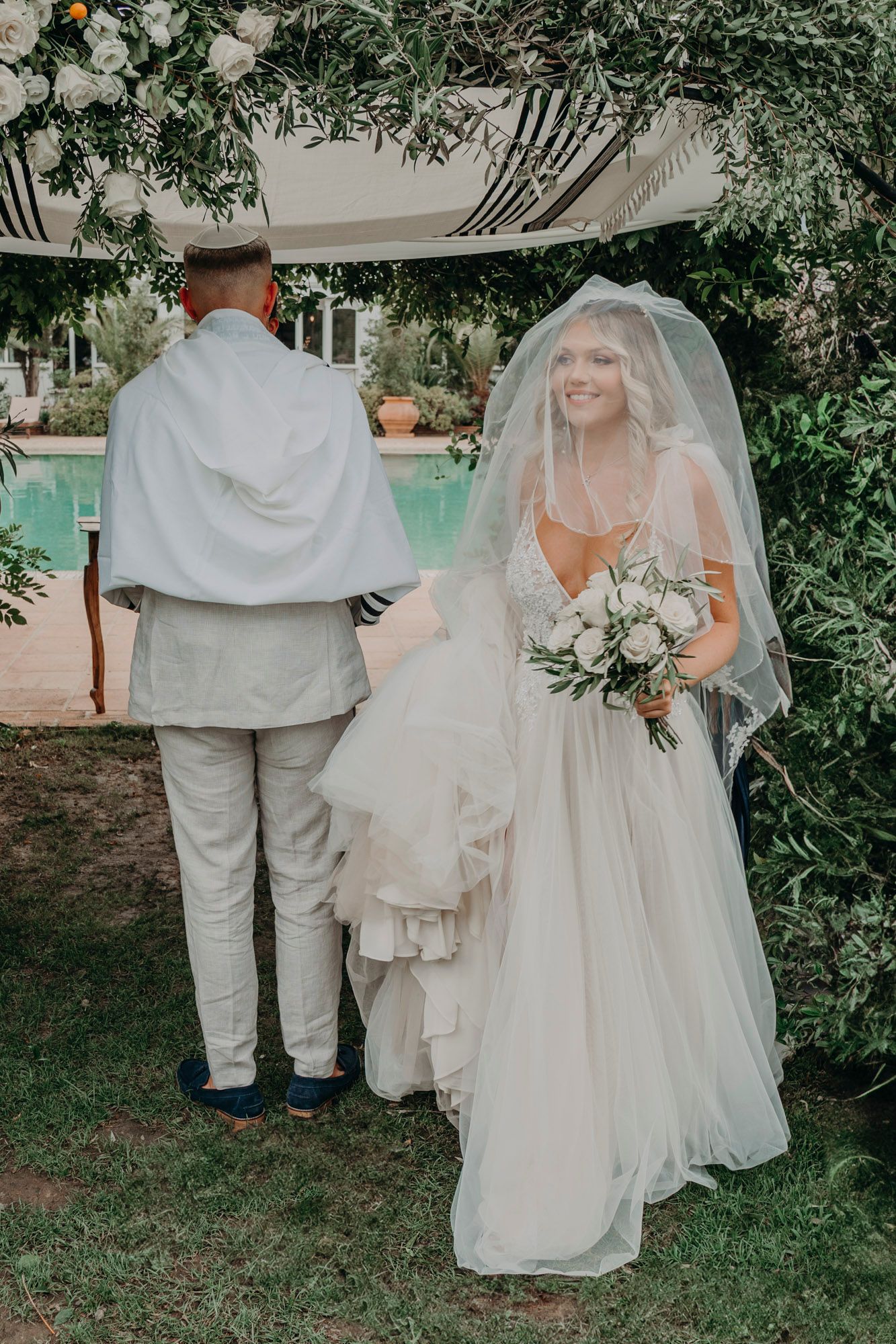 A bride in a flowing white dress stands with someone in a light suit under a garden arch near a pool for a wedding ceremony.