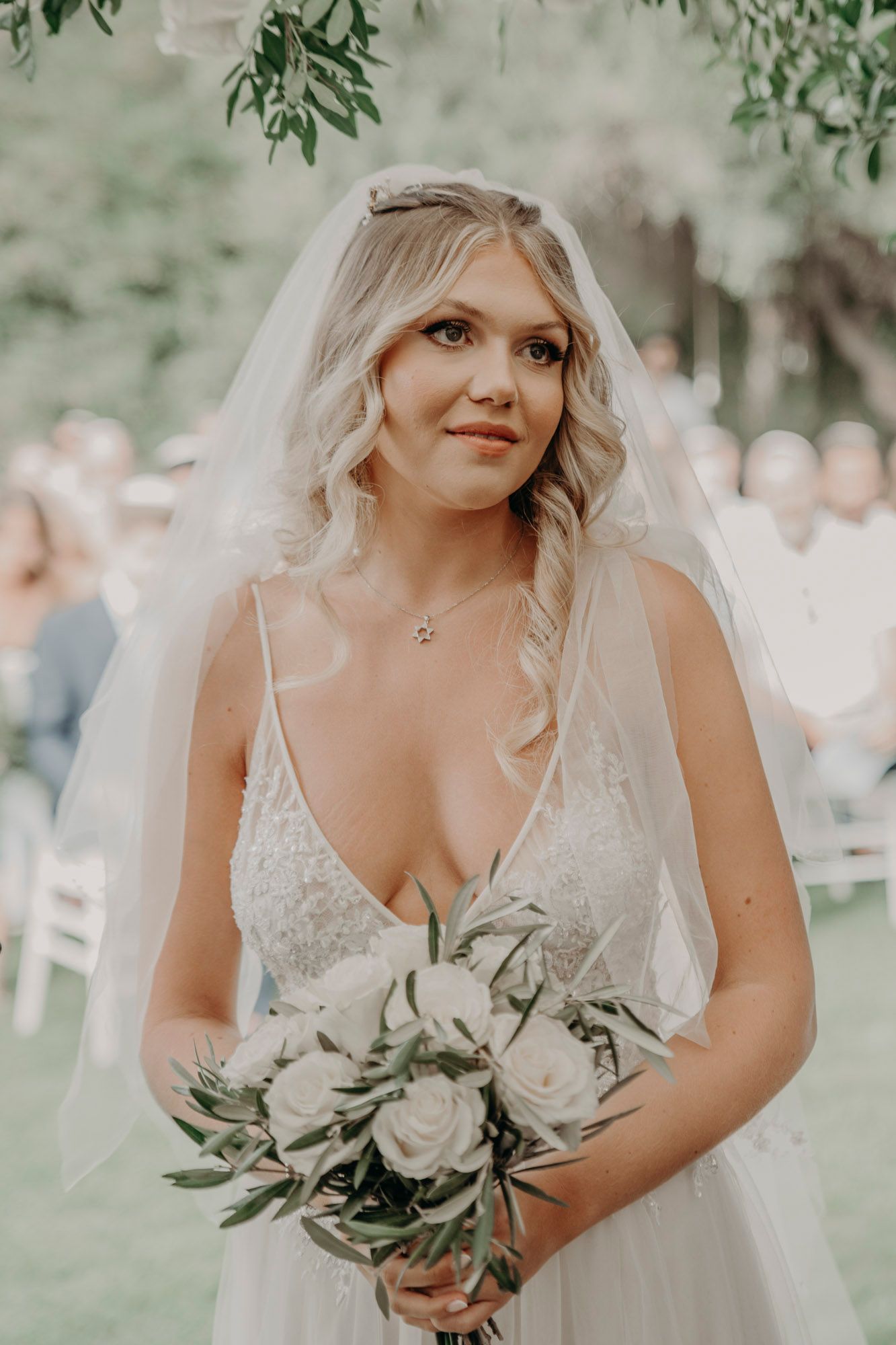 A bride in a white lace dress holds a bouquet of white roses and greenery during an outdoor wedding ceremony.