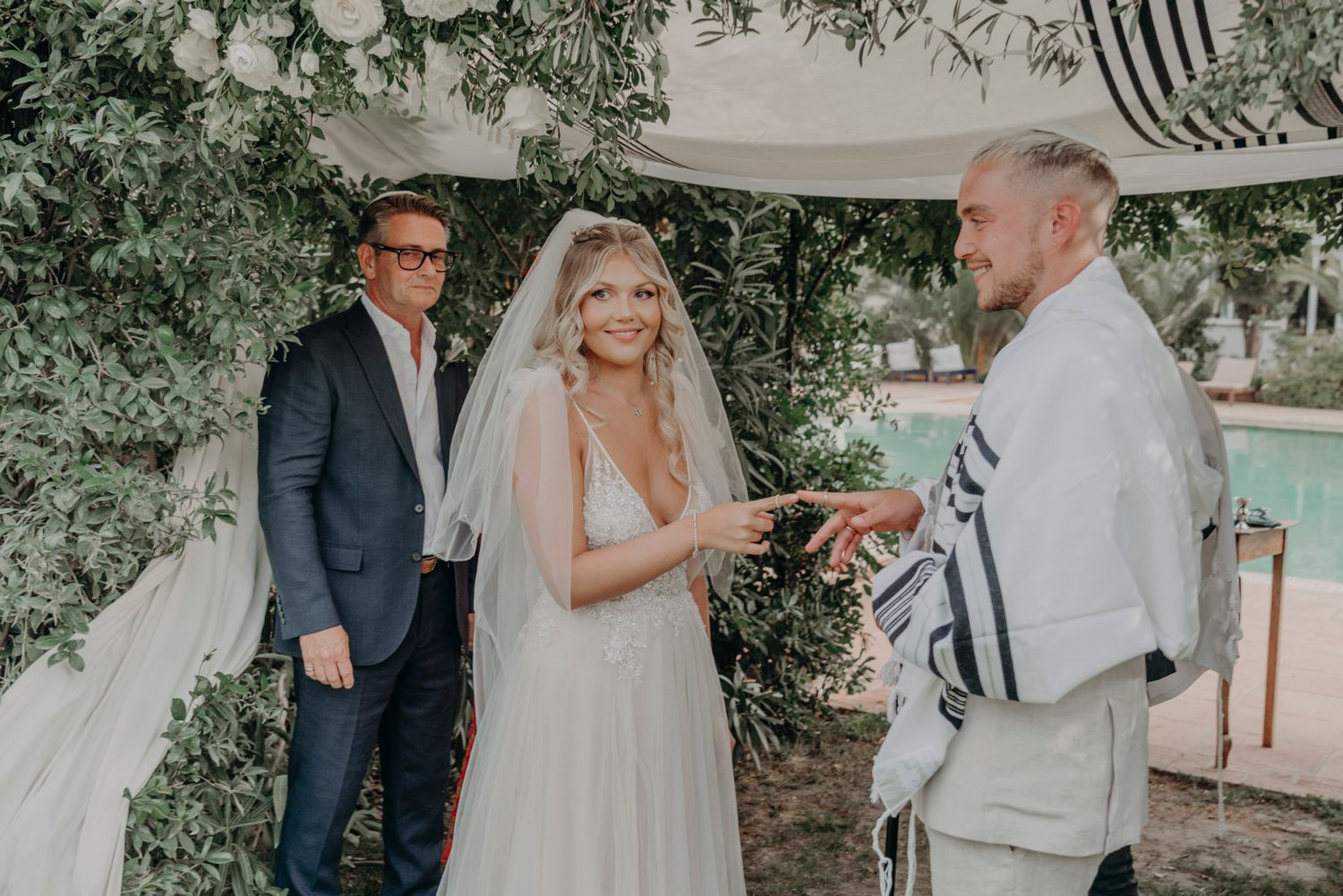 A wedding ceremony takes place outdoors under greenery with participants standing beneath white fabric draping.