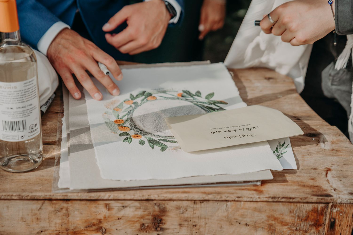 Hands resting on a rustic wooden table with a floral decorated paper invitation and glass water bottle.