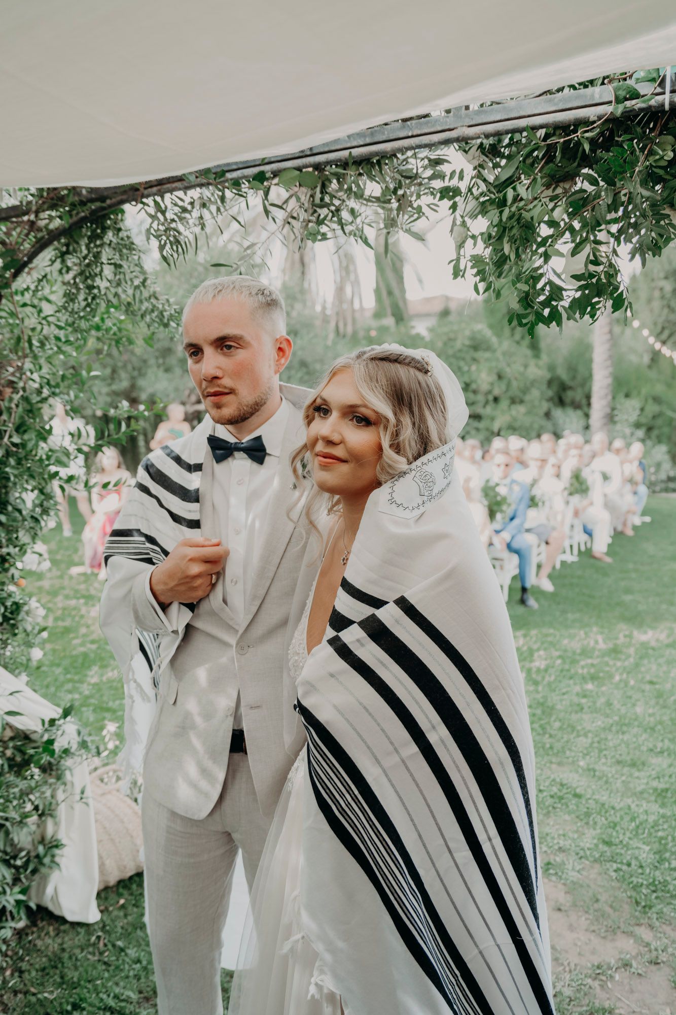 A couple wrapped in a traditional white and black striped tallit during an outdoor wedding ceremony in a garden setting.