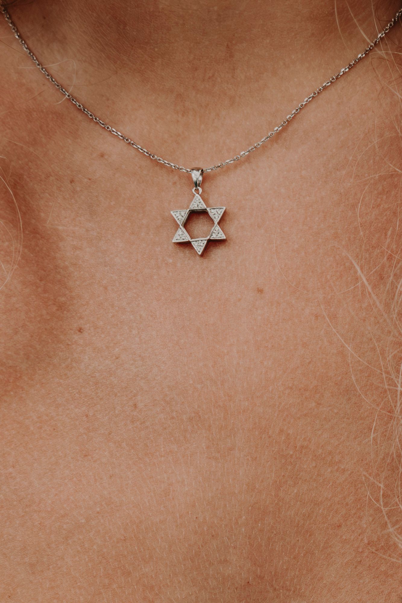 A delicate silver Star of David necklace pendant with a diamond accent shown against bare skin.