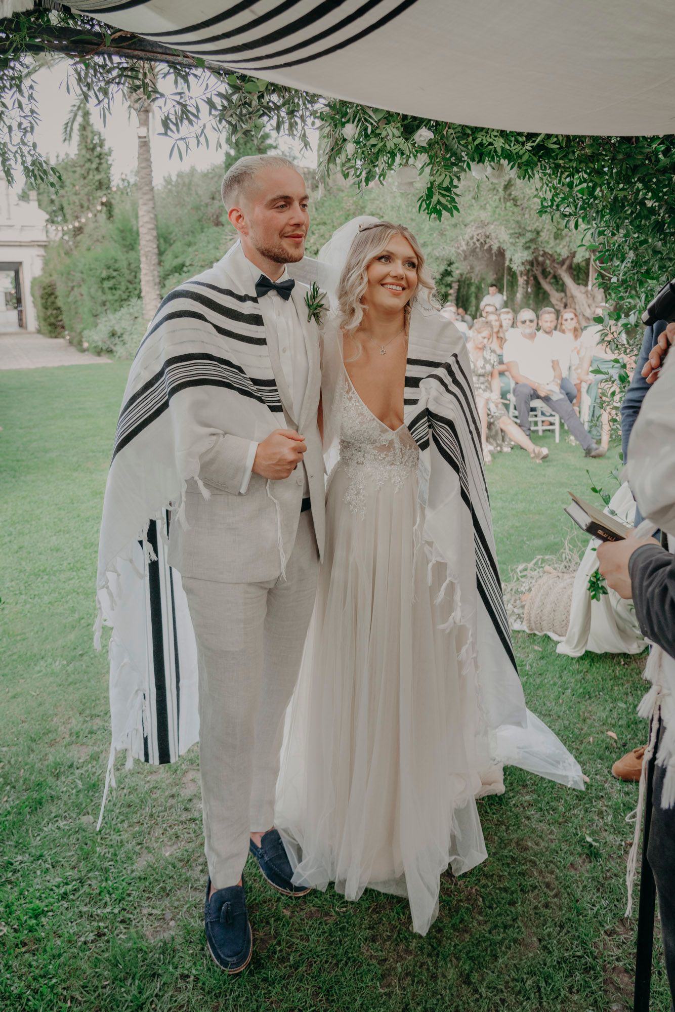 Couple stand together under outdoor ceremony canopy wearing traditional prayer shawls with bride in flowing white gown.