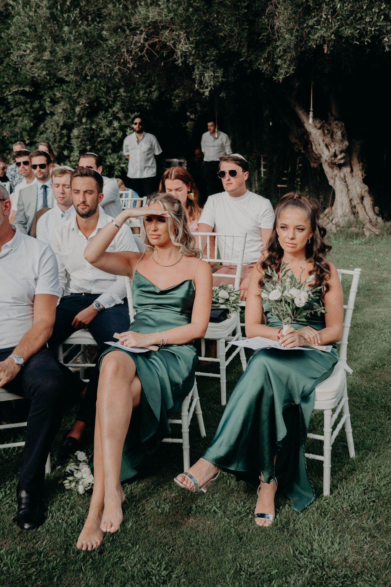 Wedding guests in formal attire seated outdoors on white chairs with women wearing elegant emerald green dresses.