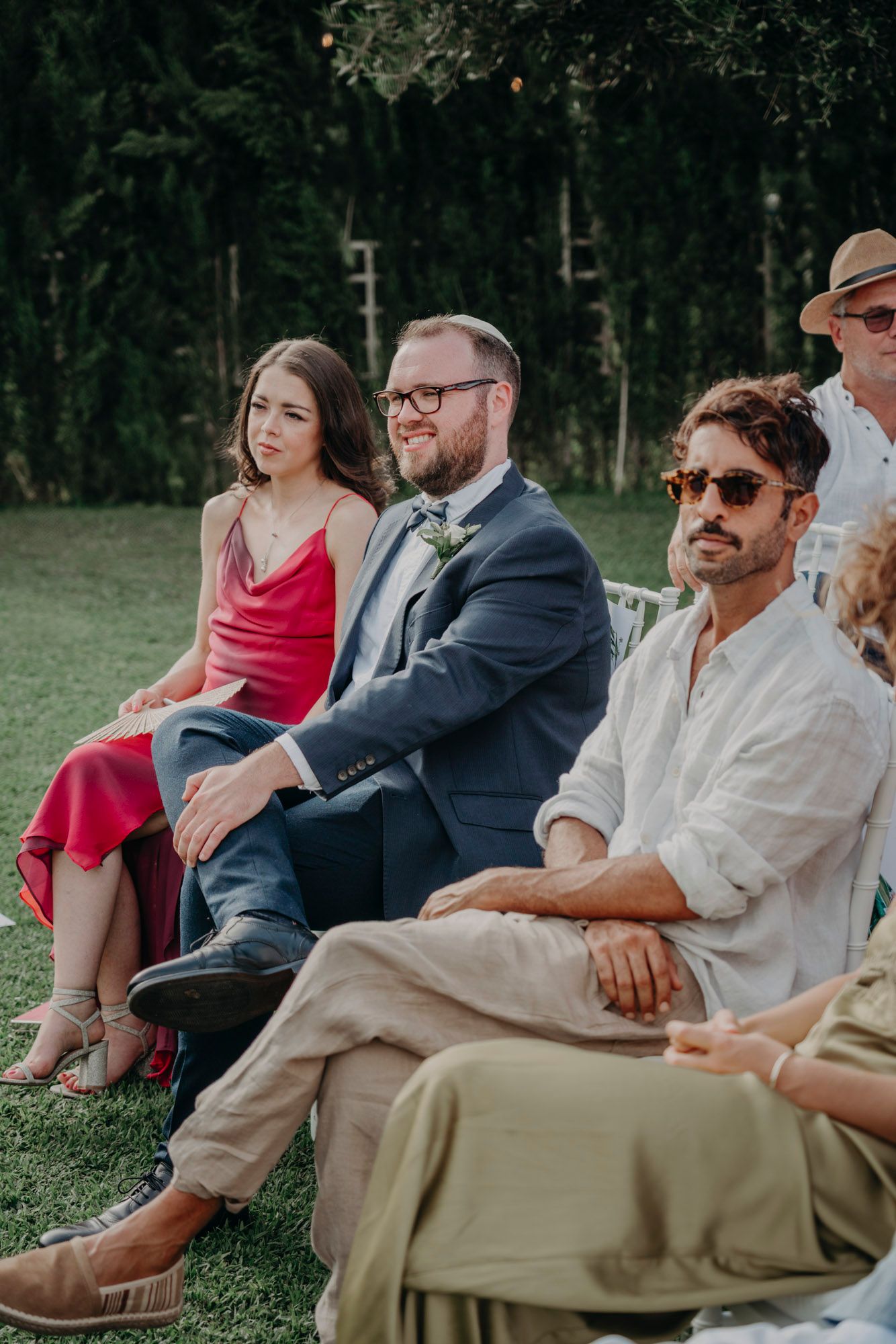 Wedding guests seated outdoors on chairs in casual and formal attire during a summer ceremony.