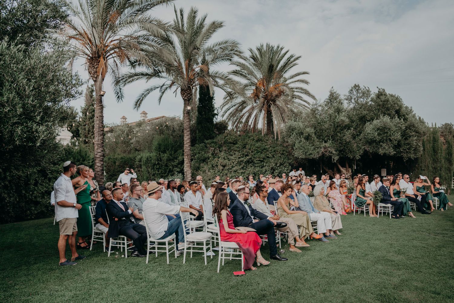 Wedding guests seated on white chairs on a lush green lawn with tall palm trees under a cloudy sky during an outdoor ceremony.