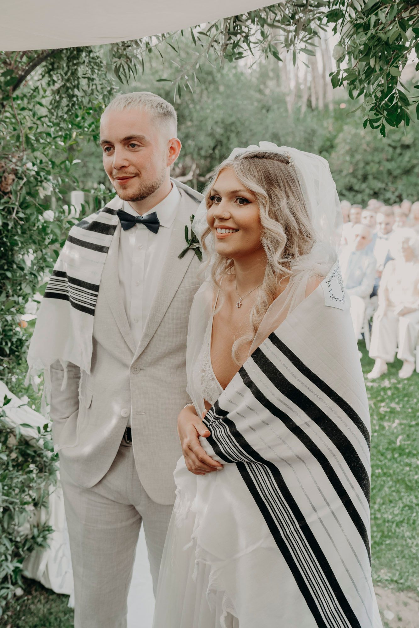 A bride and groom at their Jewish wedding ceremony wearing traditional tallit prayer shawls with black stripes.