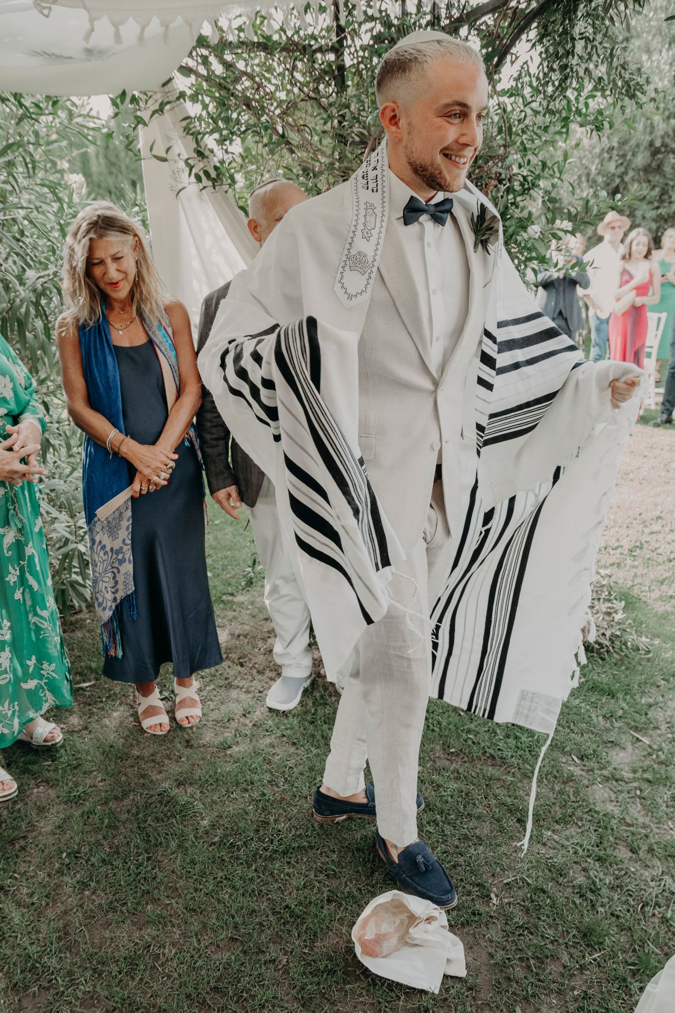 During an outdoor ceremony, a person in white formal wear and tallit stands joyfully while wearing a bow tie.