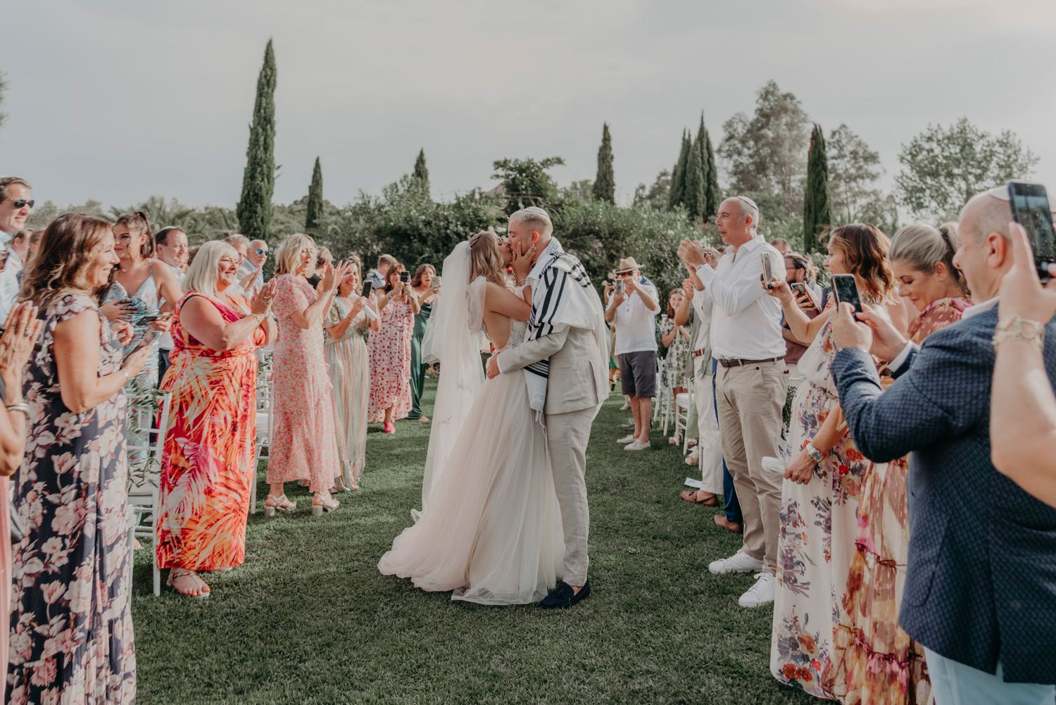 Wedding celebration on a lush lawn with cypress trees as guests in colorful floral dresses line the aisle during ceremony.