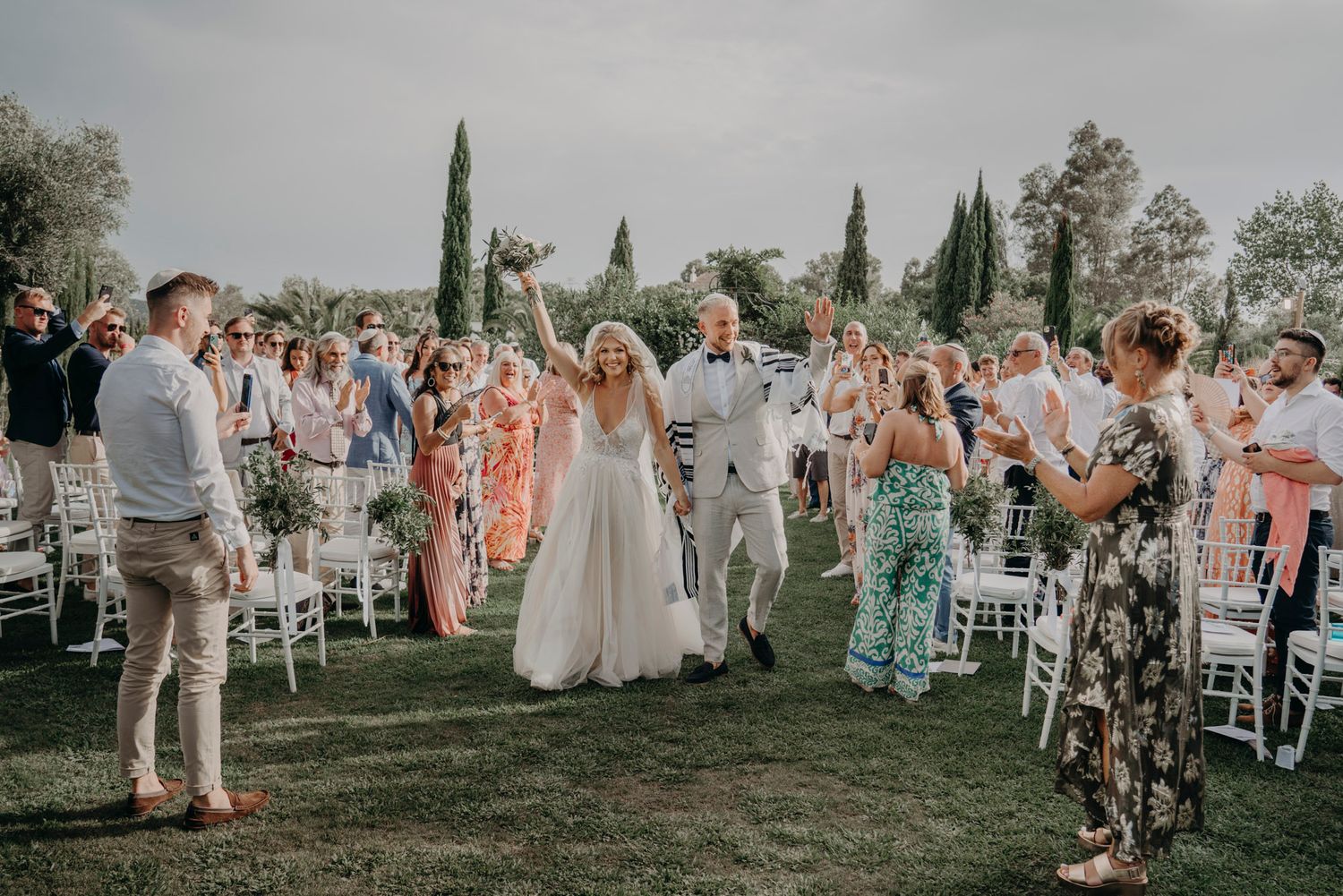 A joyful outdoor wedding celebration with guests gathered on a lawn surrounded by cypress trees during golden hour.