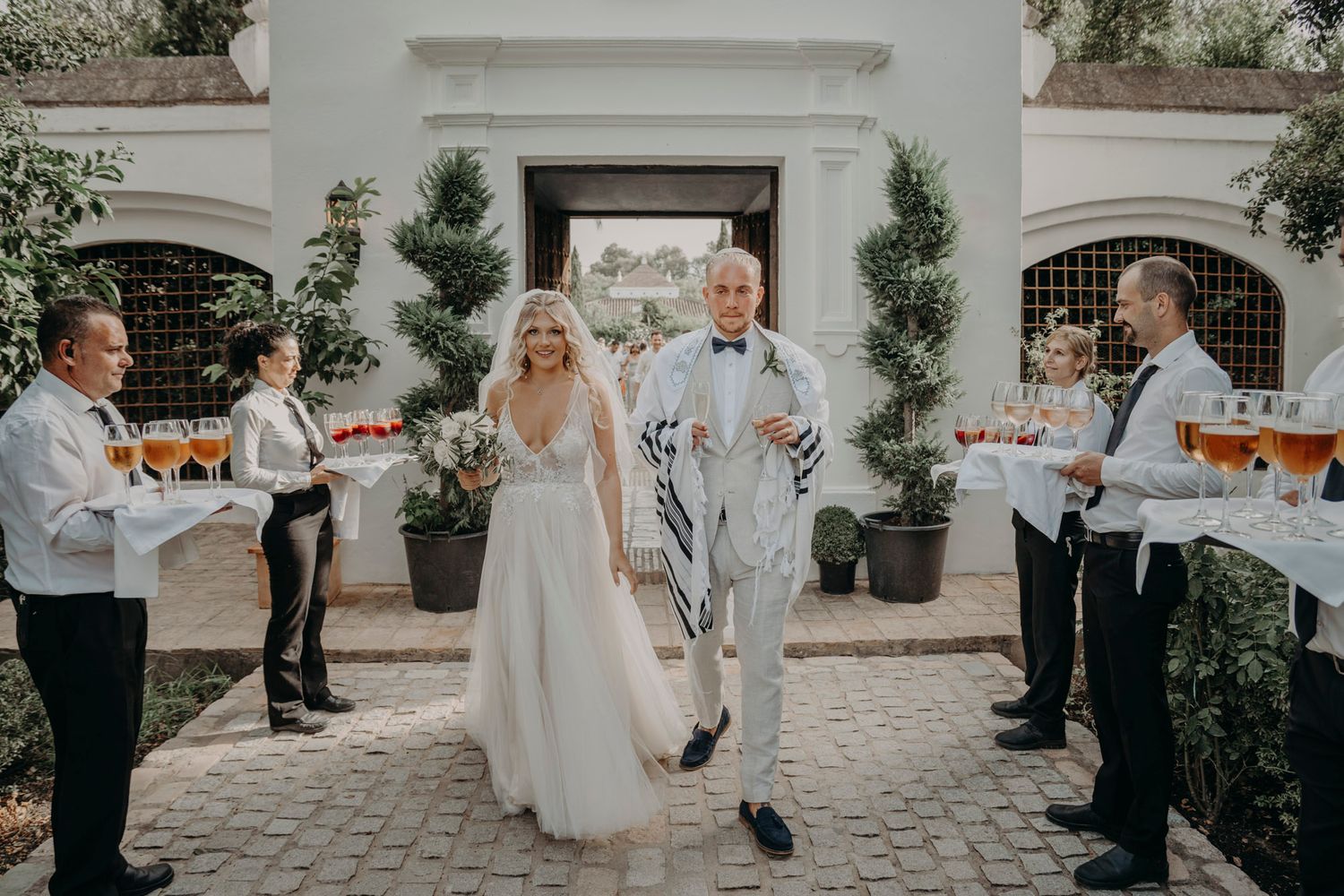 Wedding party celebrates on a paved courtyard with servers holding champagne trays as newlyweds walk together.