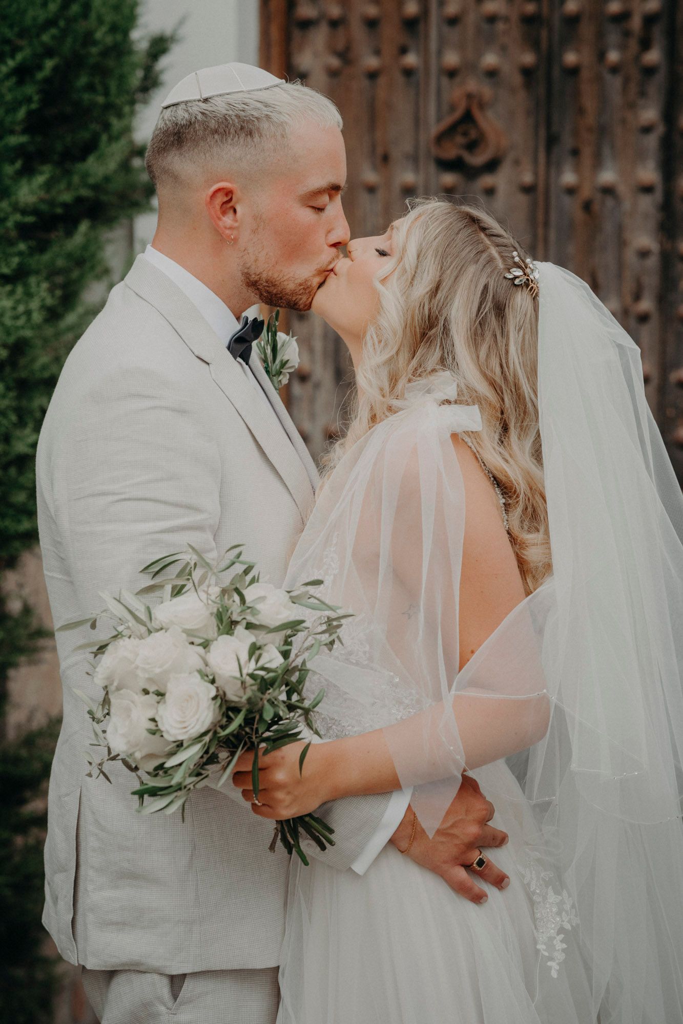 A romantic wedding couple shares a kiss while the bride holds a white floral bouquet against a rustic wooden door backdrop.
