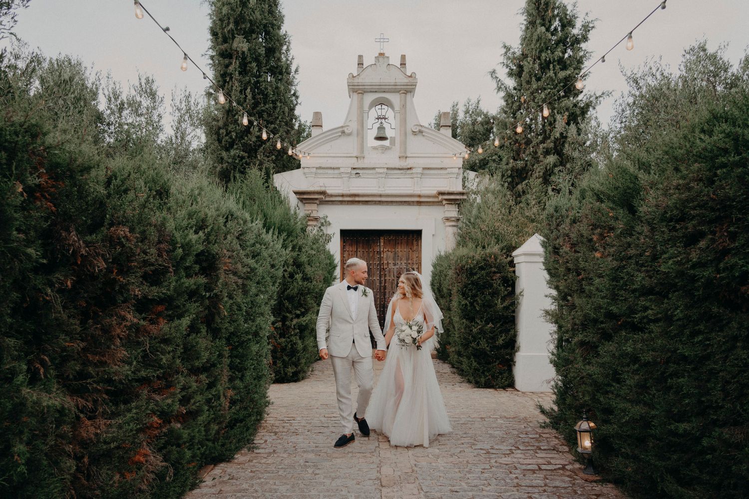 A couple walks along a garden path lined with tall hedges leading to an ornate white stone chapel entrance.