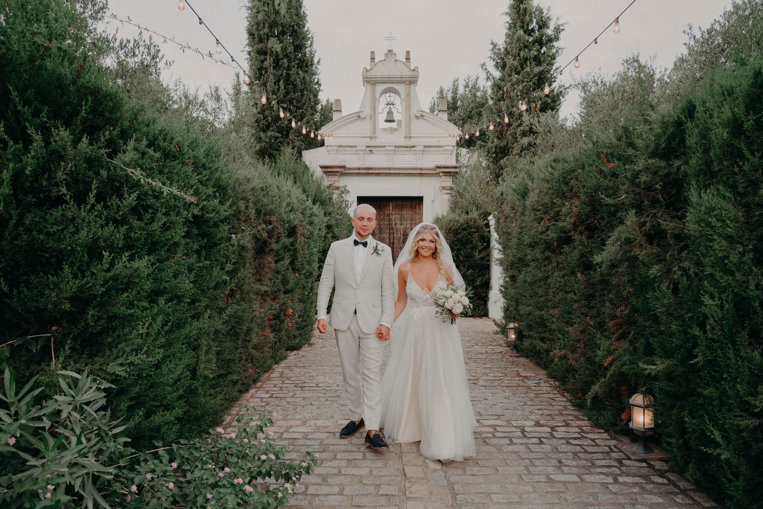 A couple walks down a stone path lined with green hedges toward an elegant stone archway entrance.