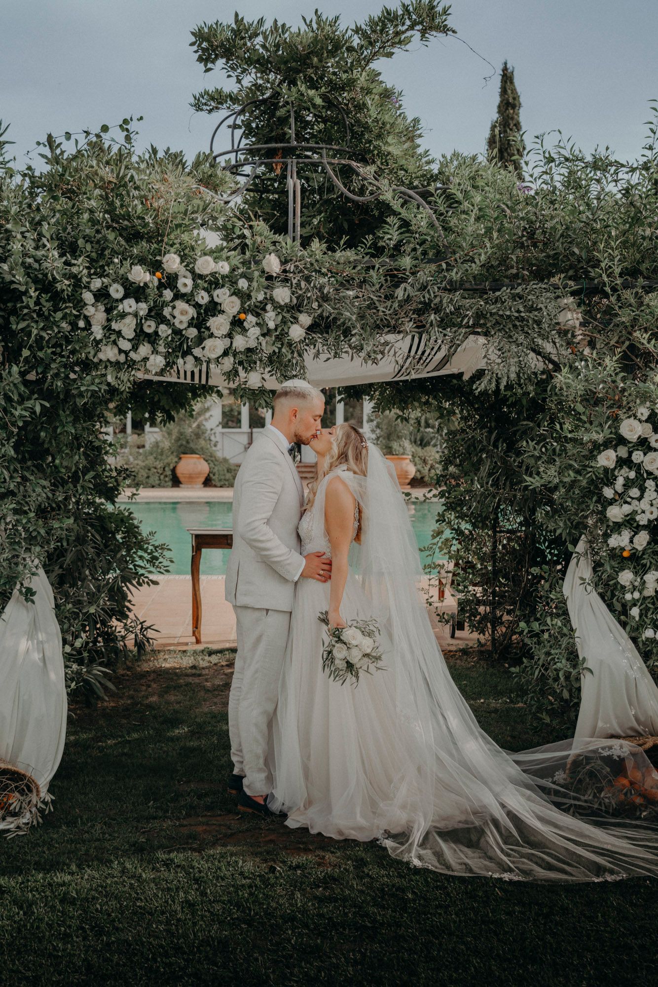 A couple shares a romantic moment under a floral arch at their outdoor garden wedding with dreamy lighting.