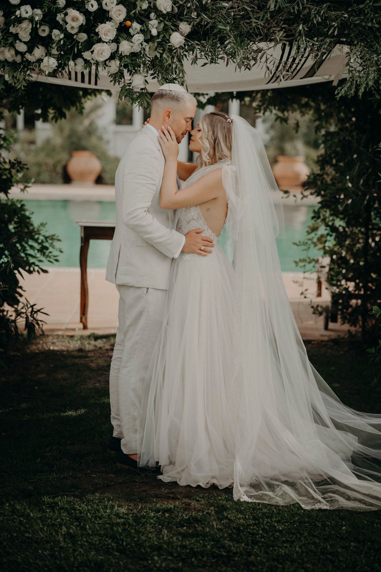 A romantic outdoor wedding moment captured beneath an elegant floral archway beside a pool.