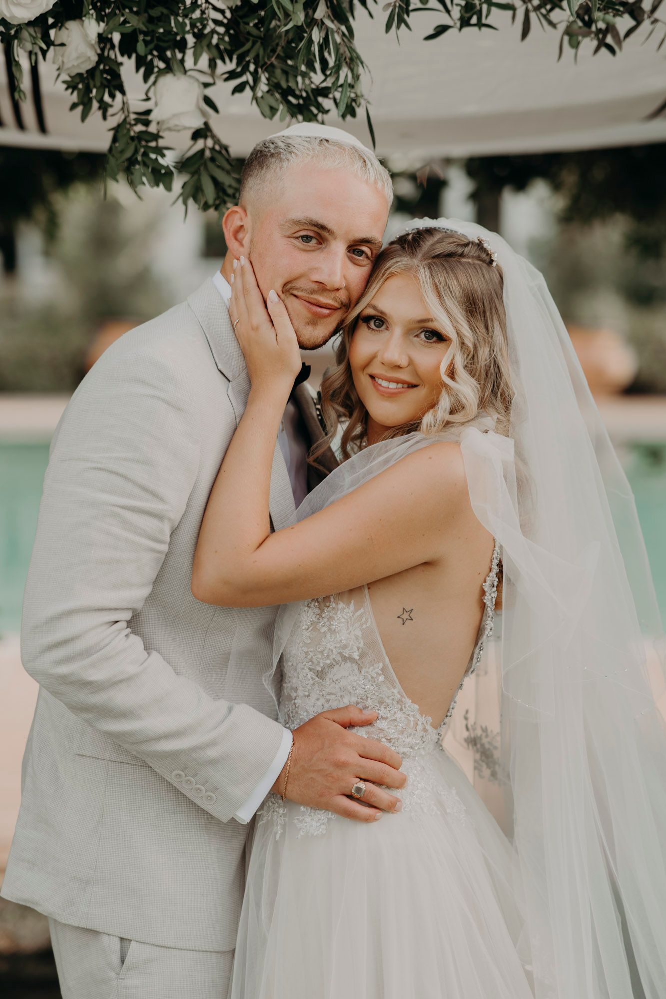 A newly married couple embraces outdoors on their wedding day wearing white attire and a flowing veil.