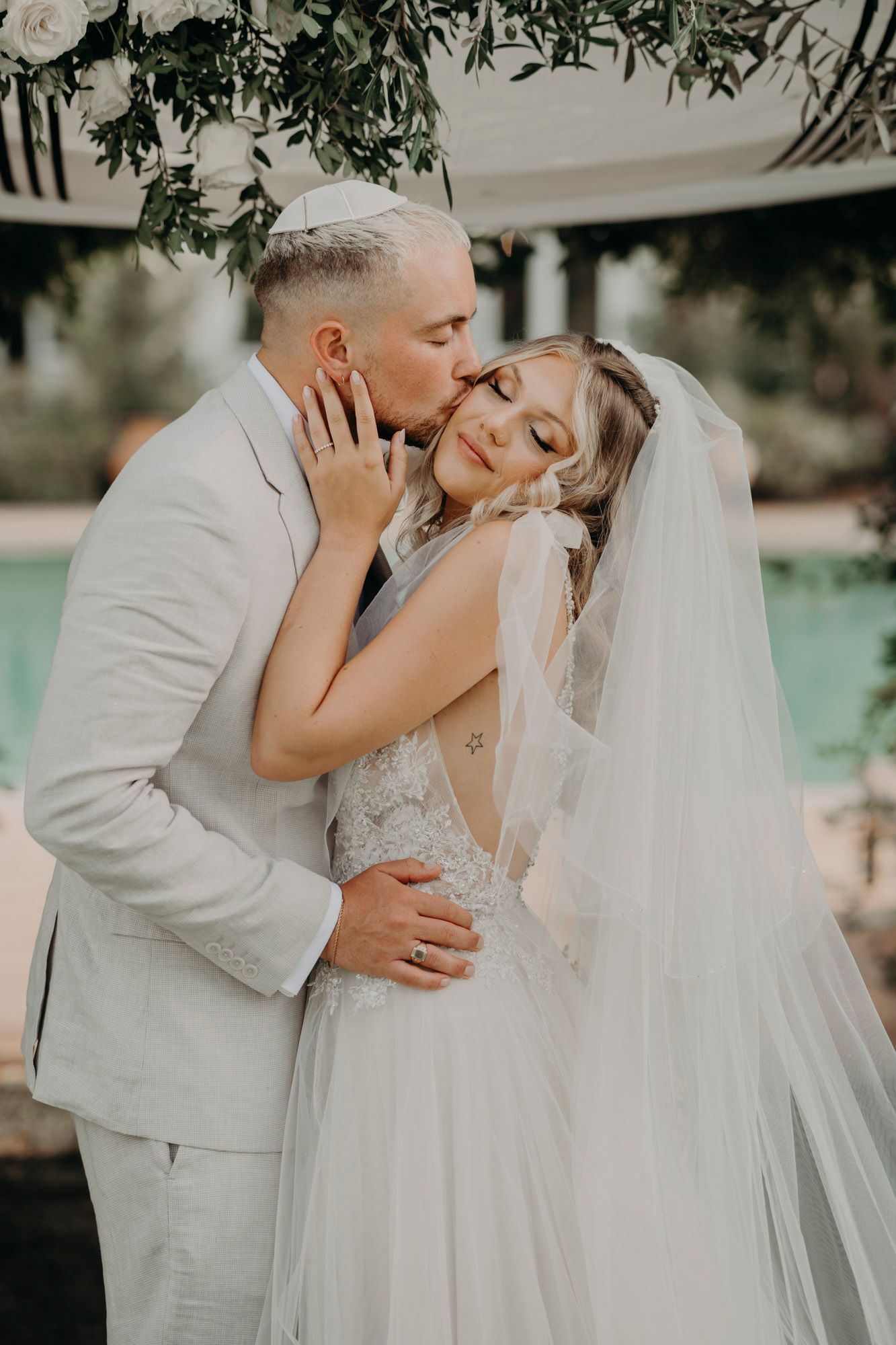 Romantic wedding couple shares an intimate moment by a pool under hanging greenery at their outdoor ceremony.