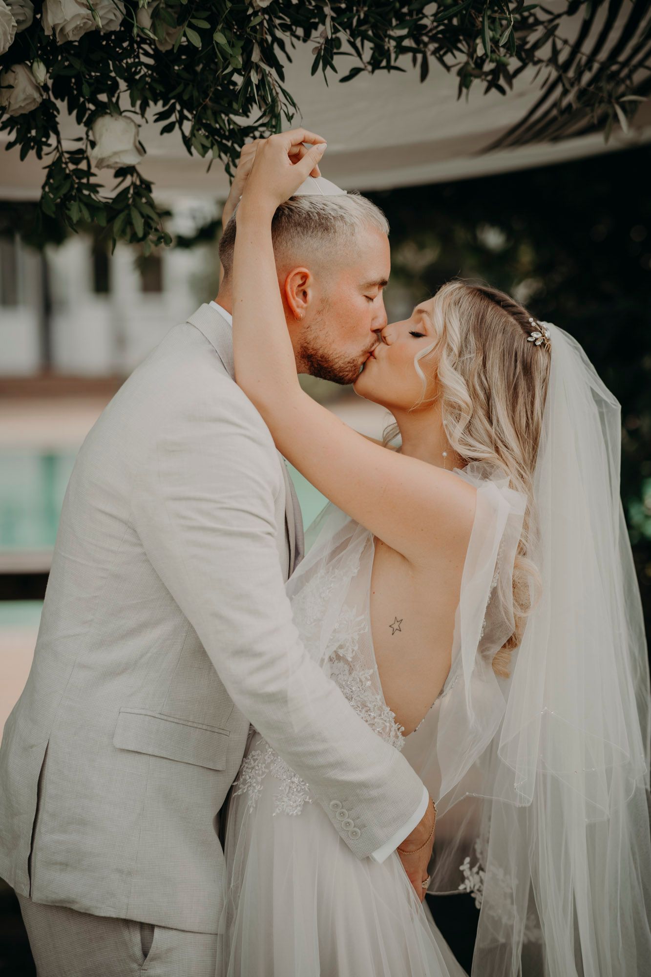 Romantic couple shares tender wedding day kiss under leafy archway at outdoor venue while wearing white attire.