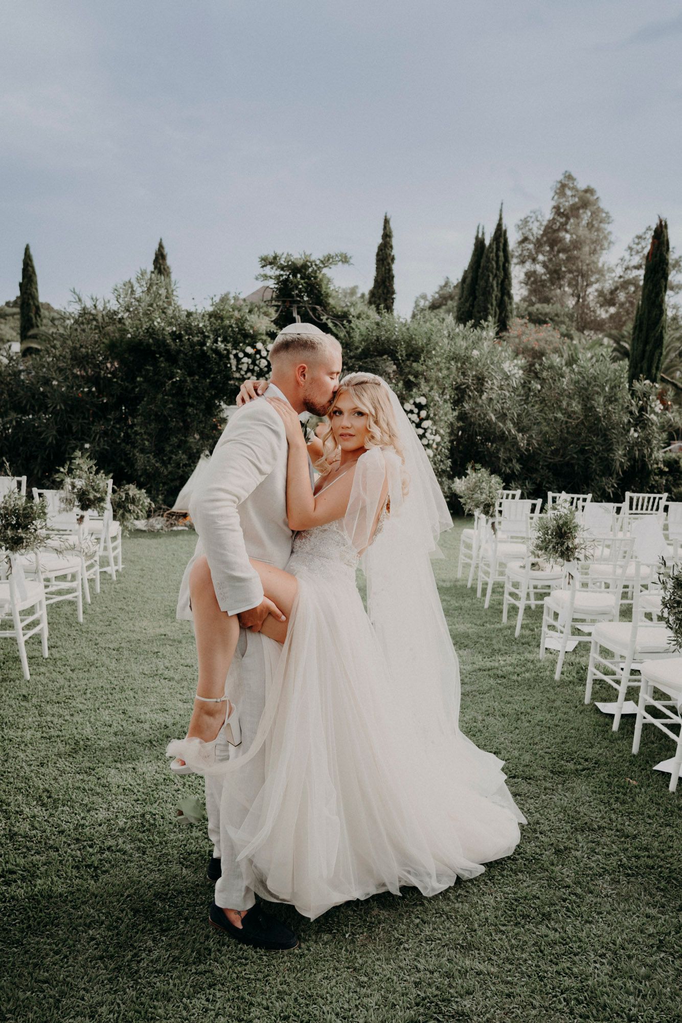 Couple shares intimate moment at outdoor garden wedding ceremony with white chairs and cypress trees in background.