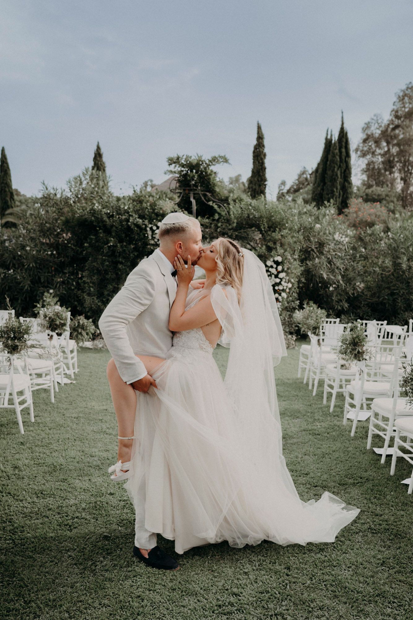 A romantic wedding moment captured in a garden setting with white chairs and cypress trees in the background.