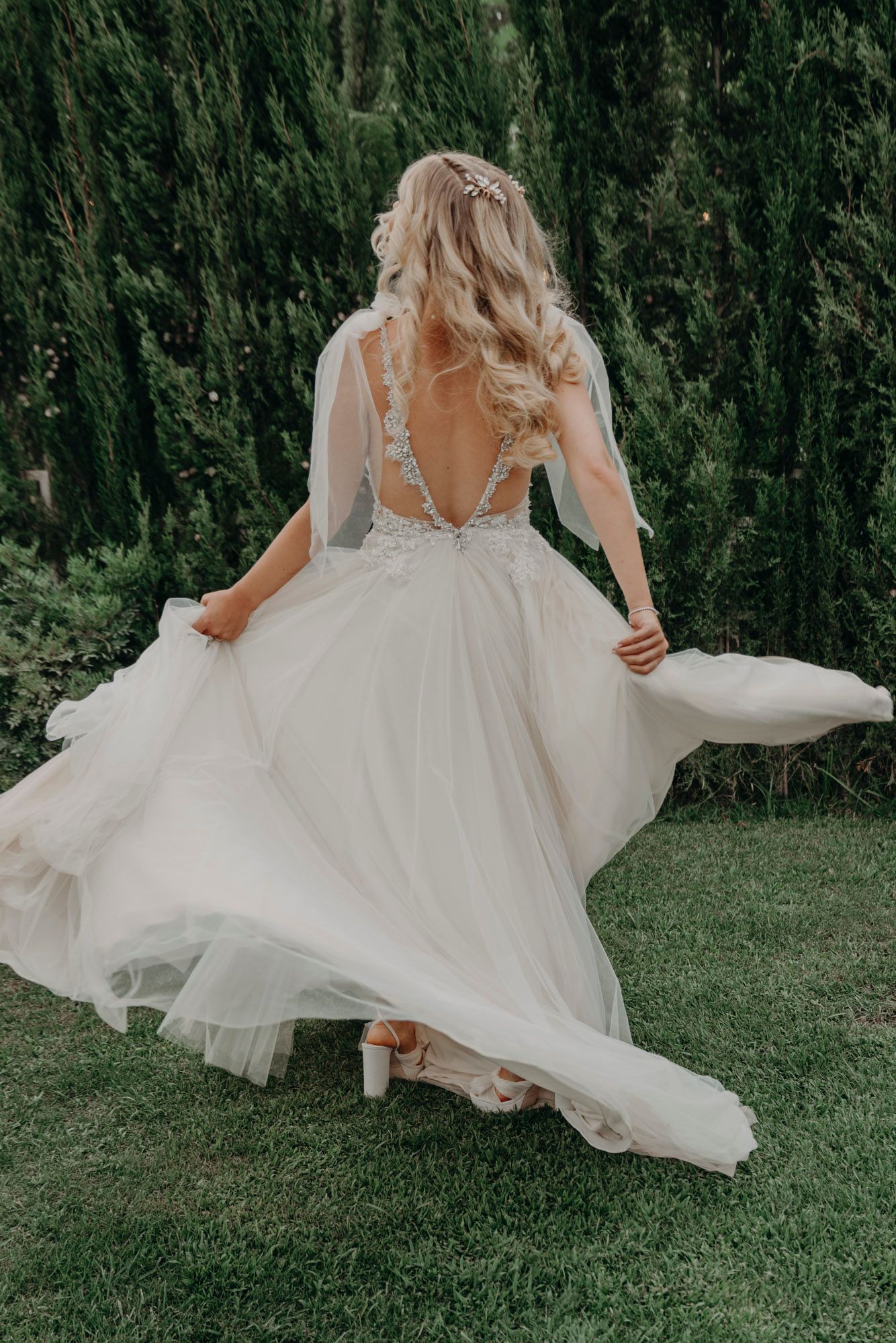 Back view of bride in flowing white tulle gown twirling on grass with greenery backdrop.