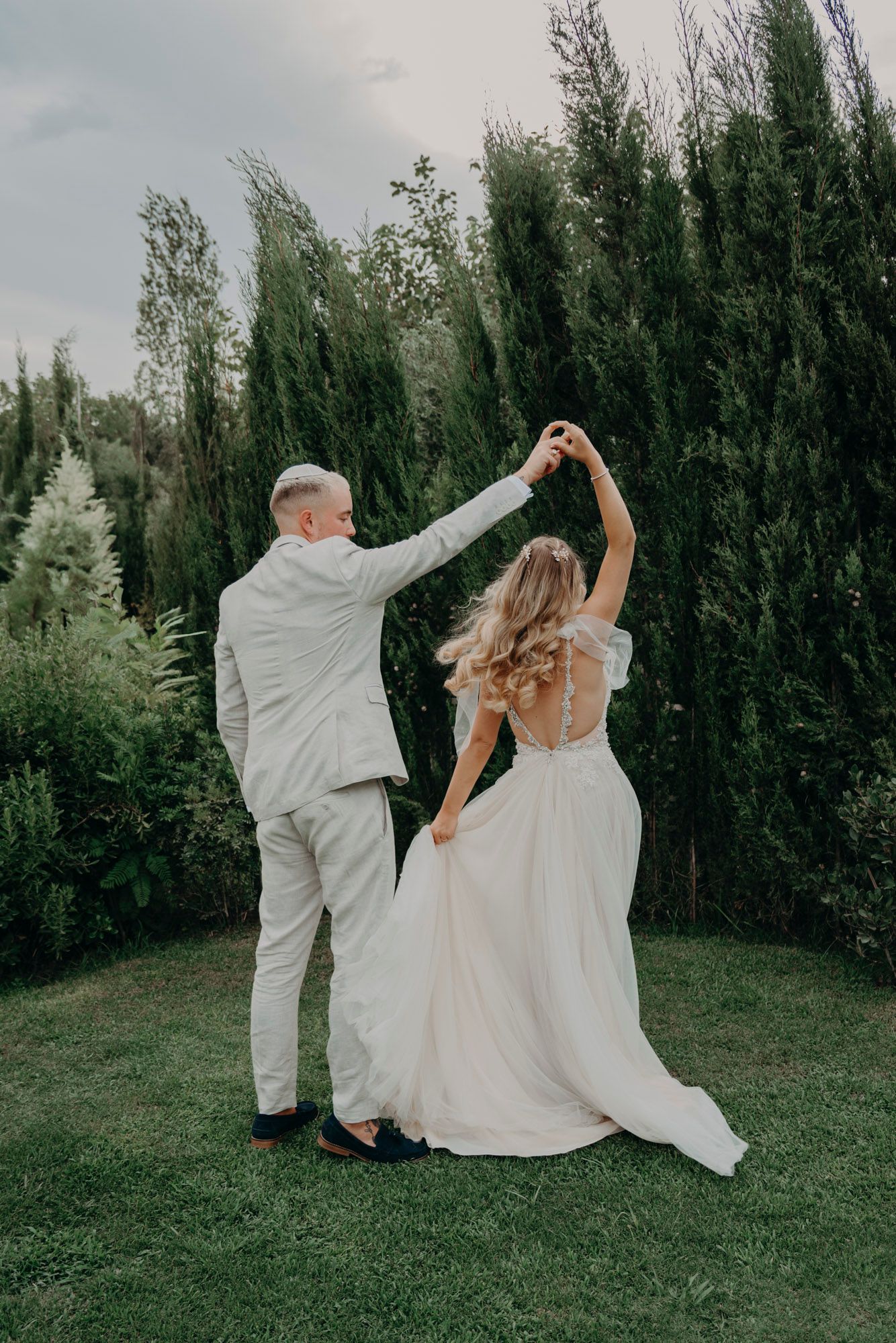 A couple in wedding attire dancing together on a green lawn with tall cypress trees in the background.