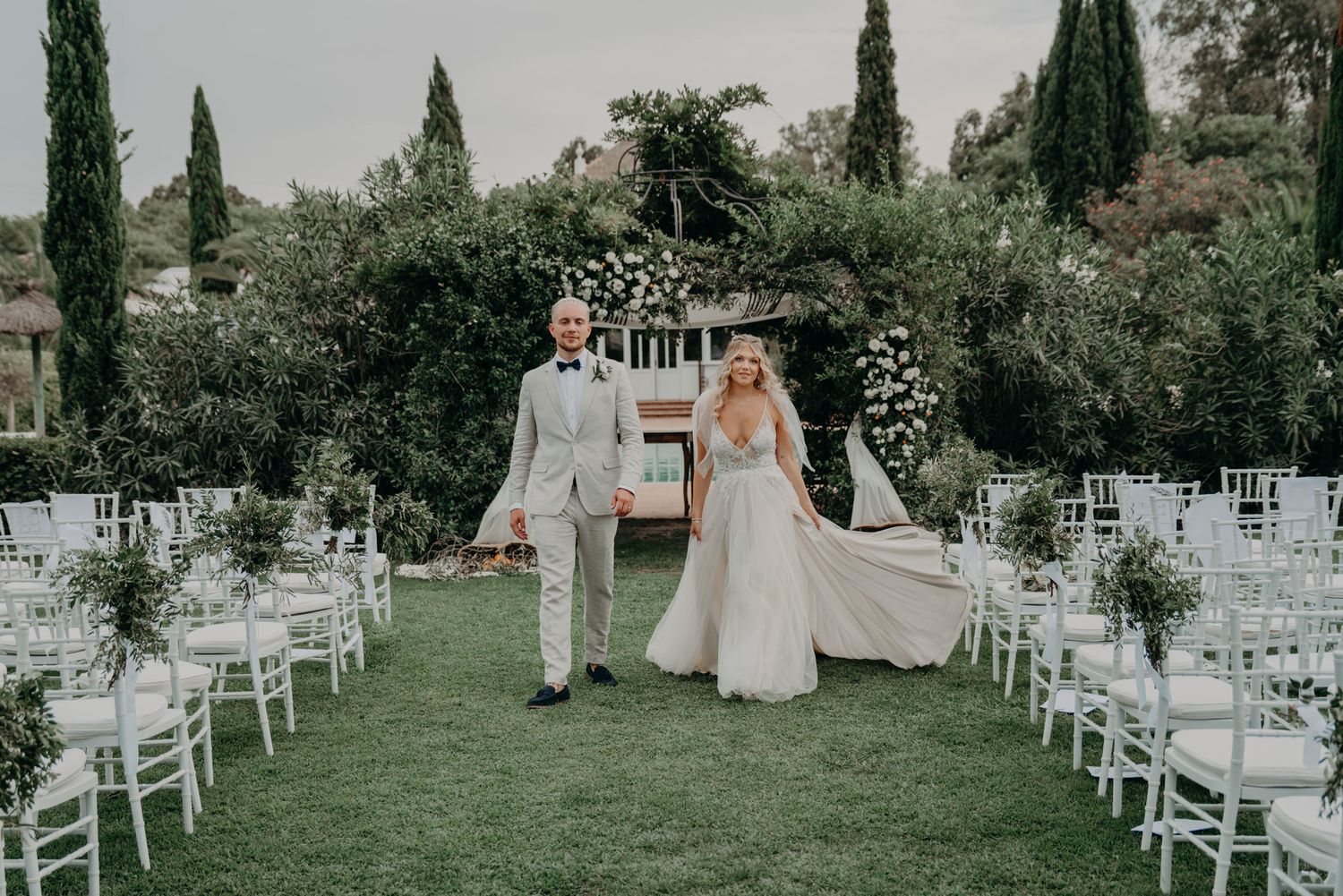 A couple walks down a grassy aisle between white chairs at an outdoor wedding ceremony with cypress trees in background.