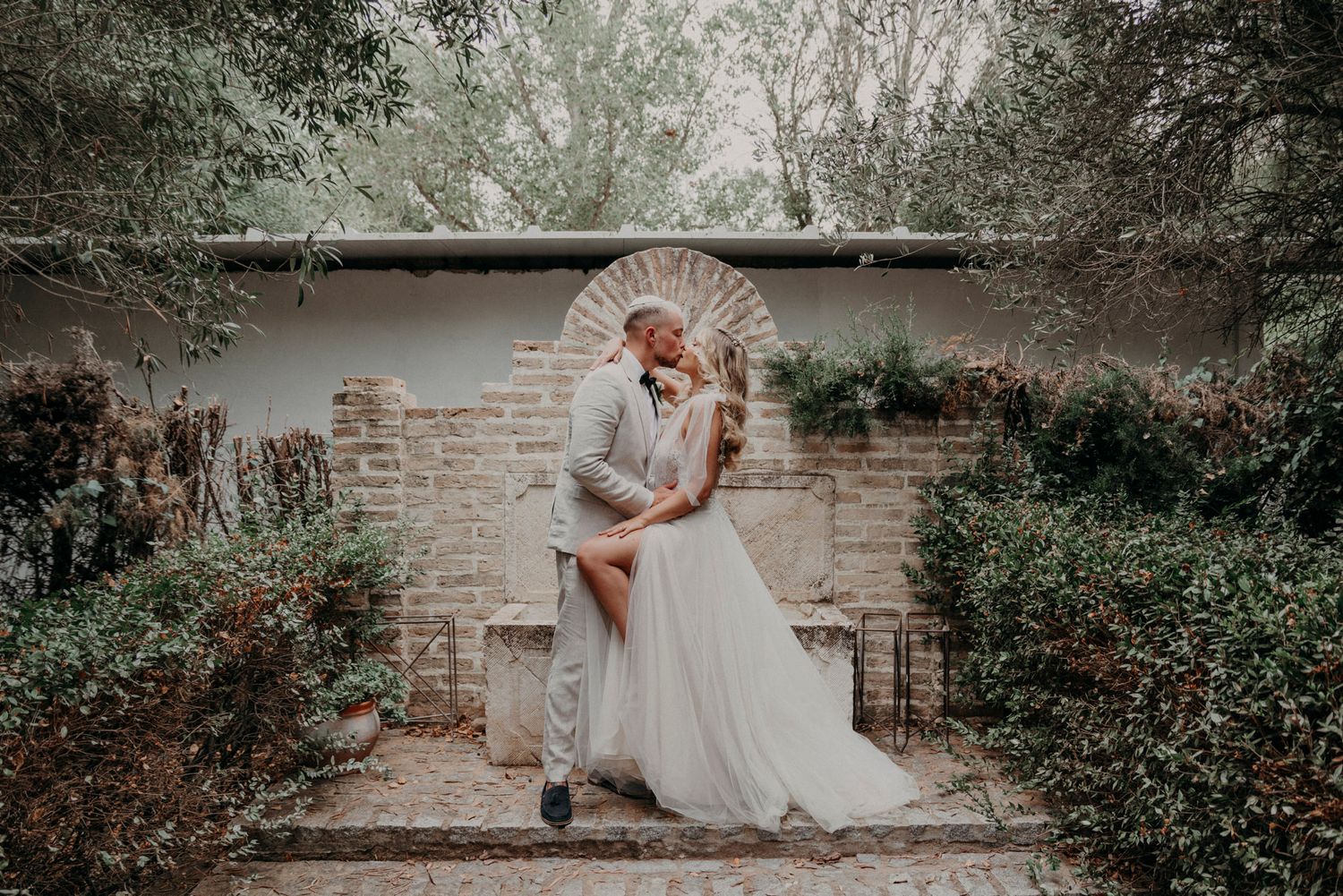 A romantic wedding couple embraces in a garden setting with brick wall and lush greenery backdrop.