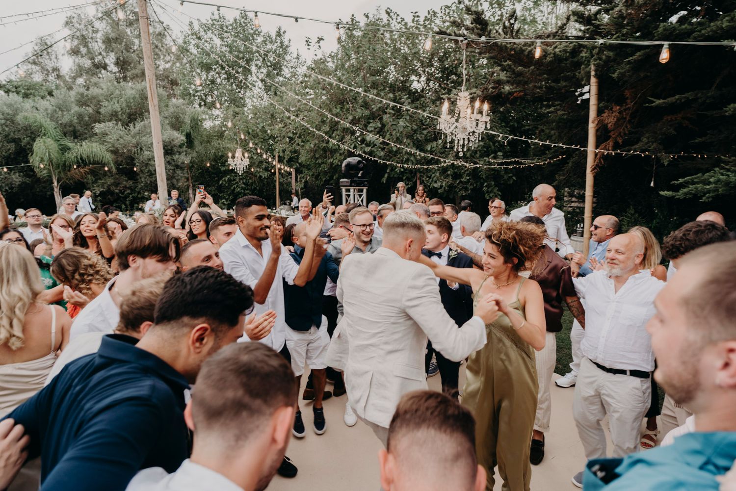 A large group of wedding guests dance together at an outdoor reception celebration during daytime.