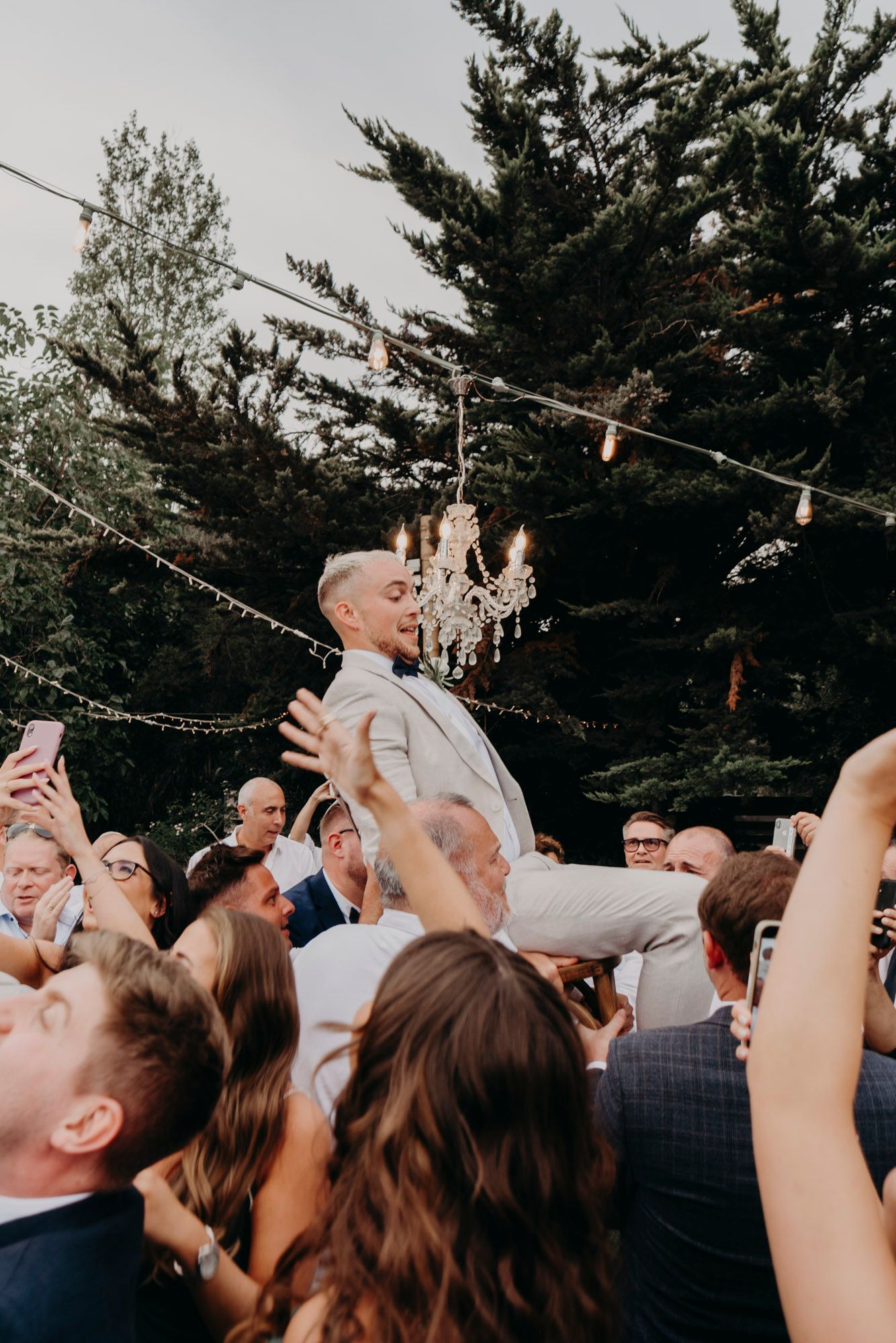 A crowd of wedding guests dance and celebrate outdoors under string lights and evergreen trees at dusk.