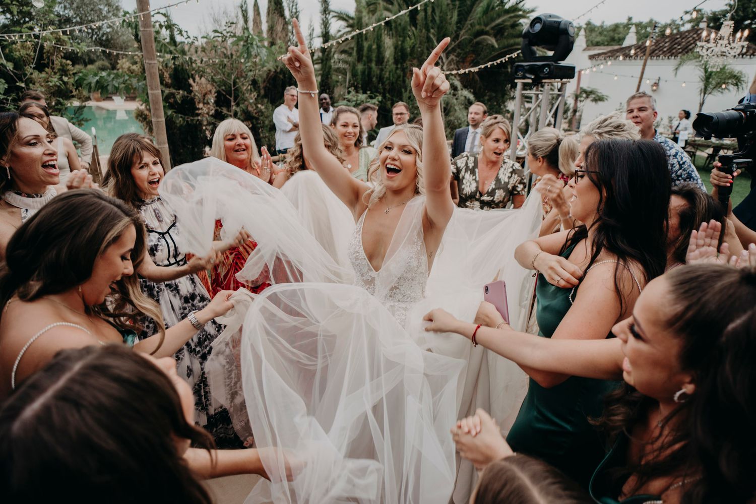 Joyful wedding celebration with guests cheering around the bride in a flowing white dress in an outdoor garden setting.