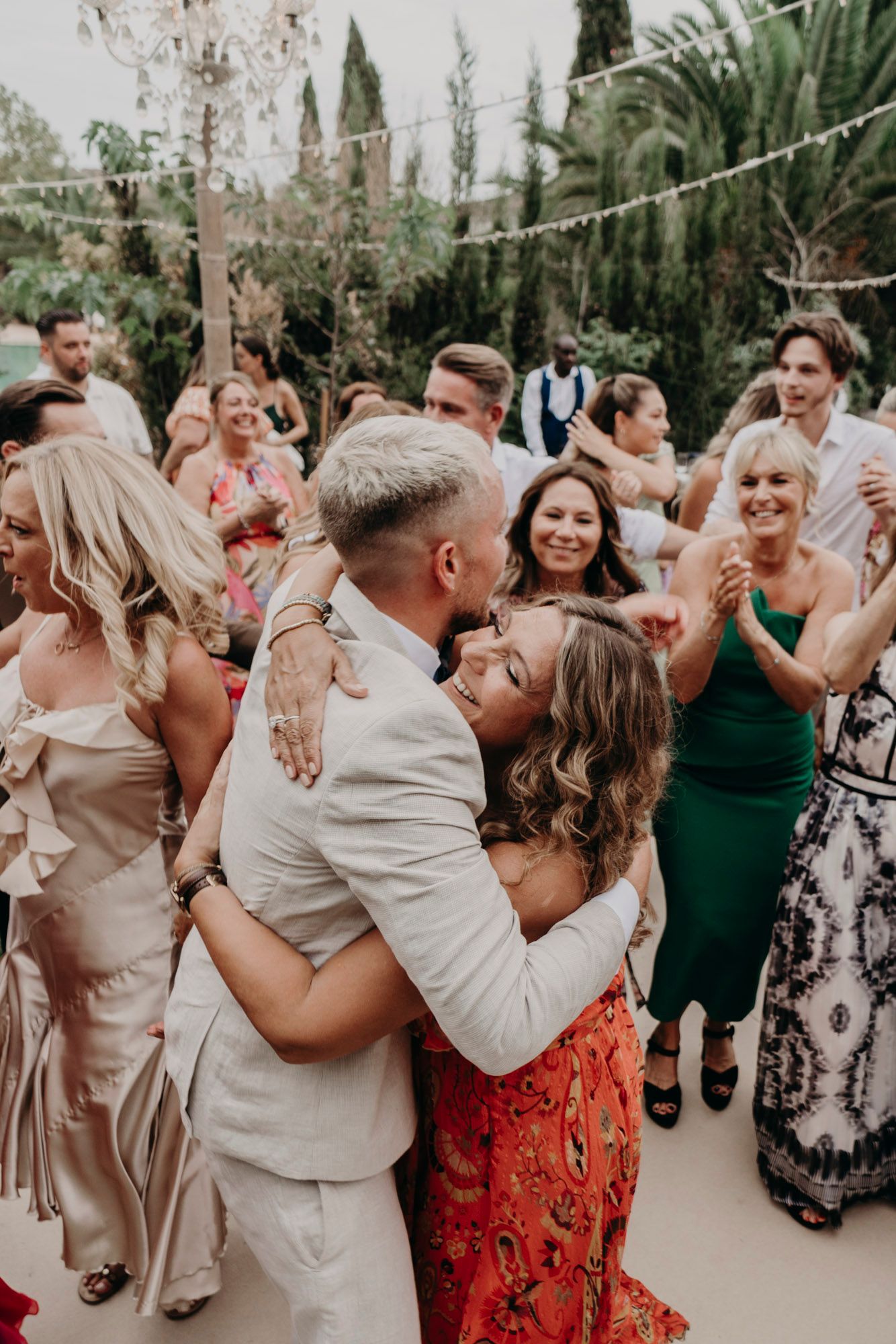 Wedding guests dance and celebrate at an outdoor reception surrounded by cypress trees and string lights.