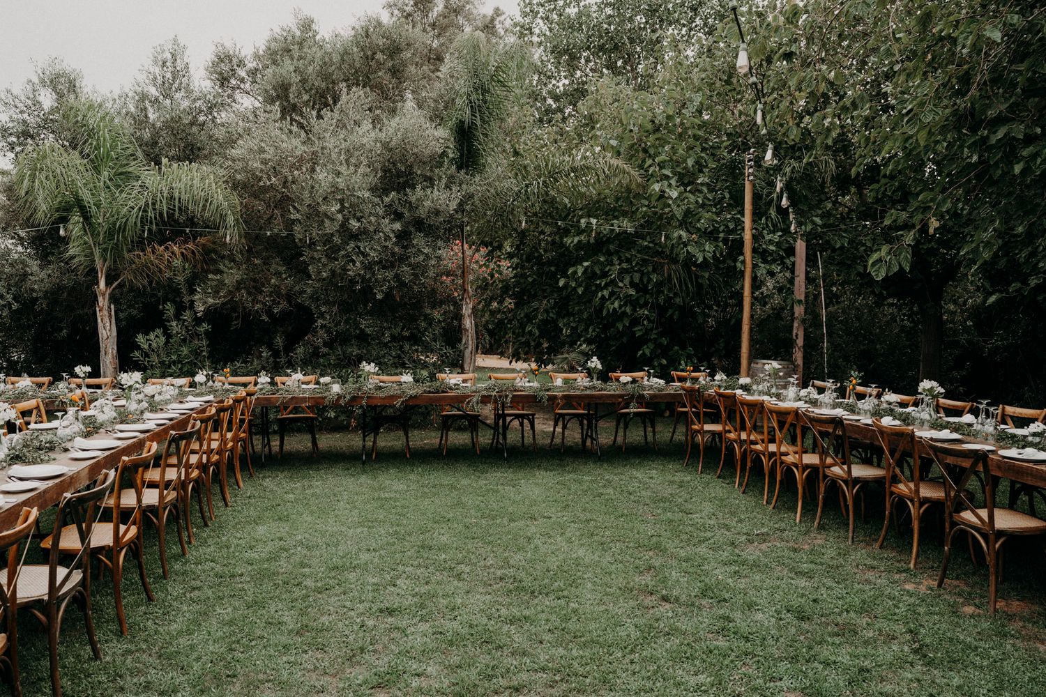 Rustic outdoor wedding reception setup with long wooden tables arranged in a U-shape on a grassy lawn surrounded by trees.
