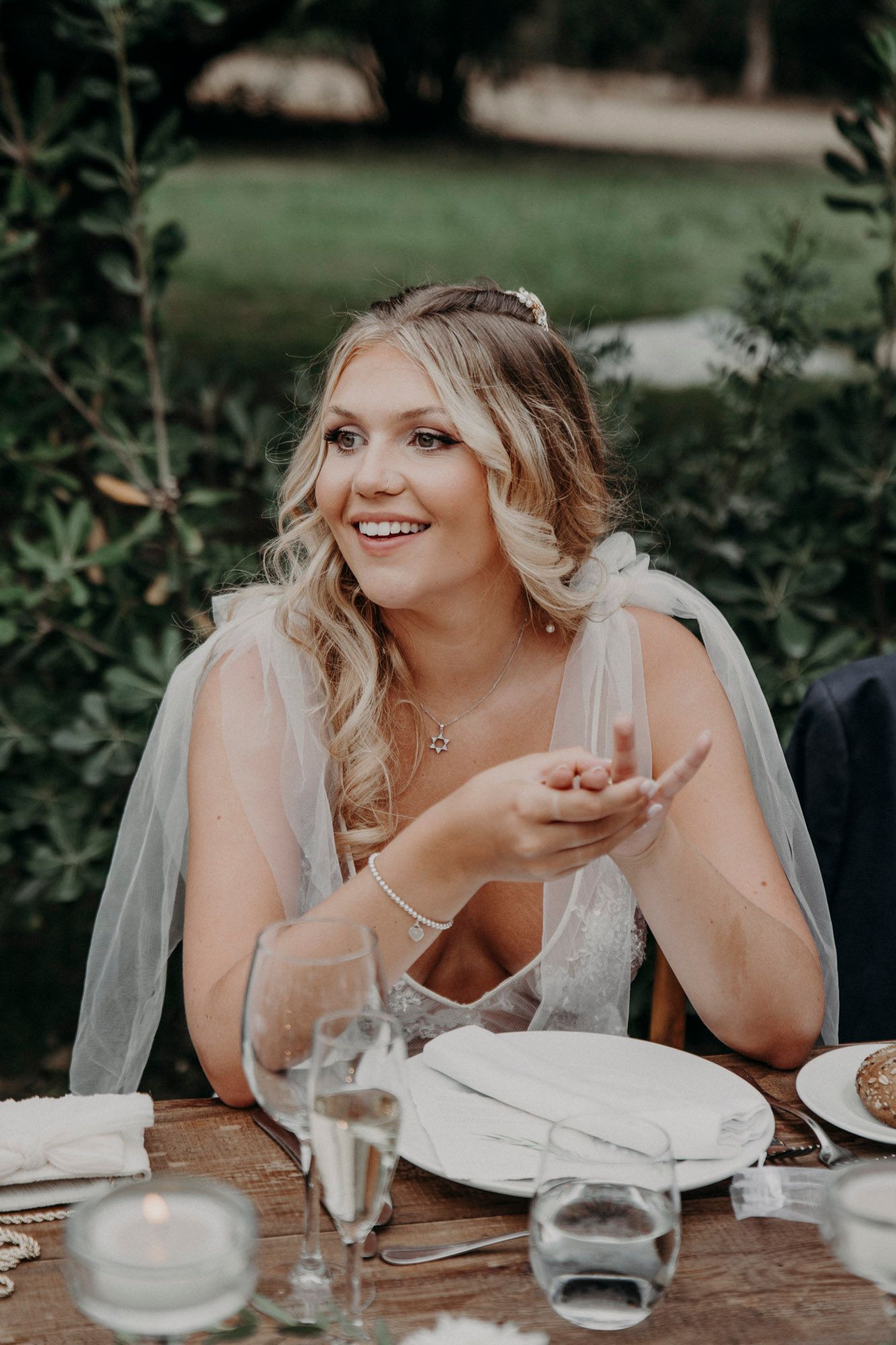 A bride smiles and gestures while seated at a table during an outdoor wedding reception.