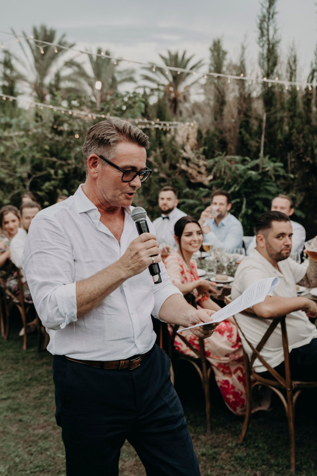 A guest giving a speech with a microphone during an outdoor garden reception with palm trees in the background.