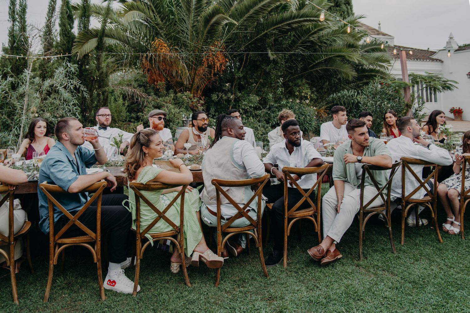 An elegant outdoor wedding reception with guests seated at long tables under palm trees in a garden setting.