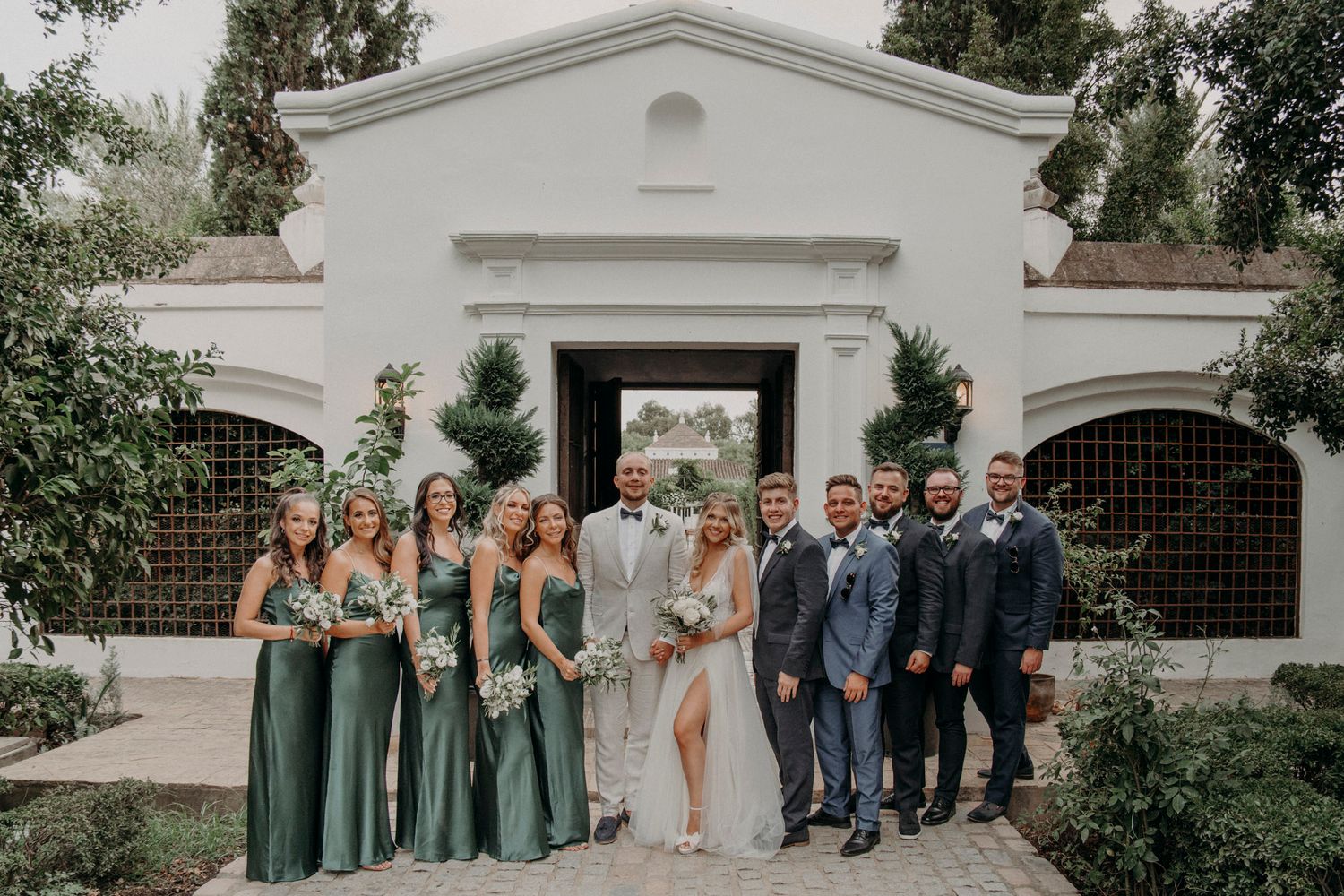 Wedding party in elegant attire poses outside white Spanish-style building with green bridesmaids dresses and blue suits.