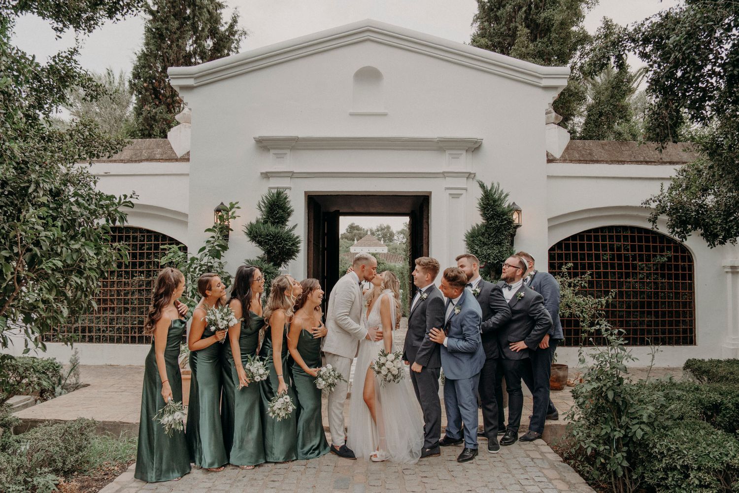 Wedding party in emerald green dresses and blue suits pose together outside white Spanish-style building entrance.